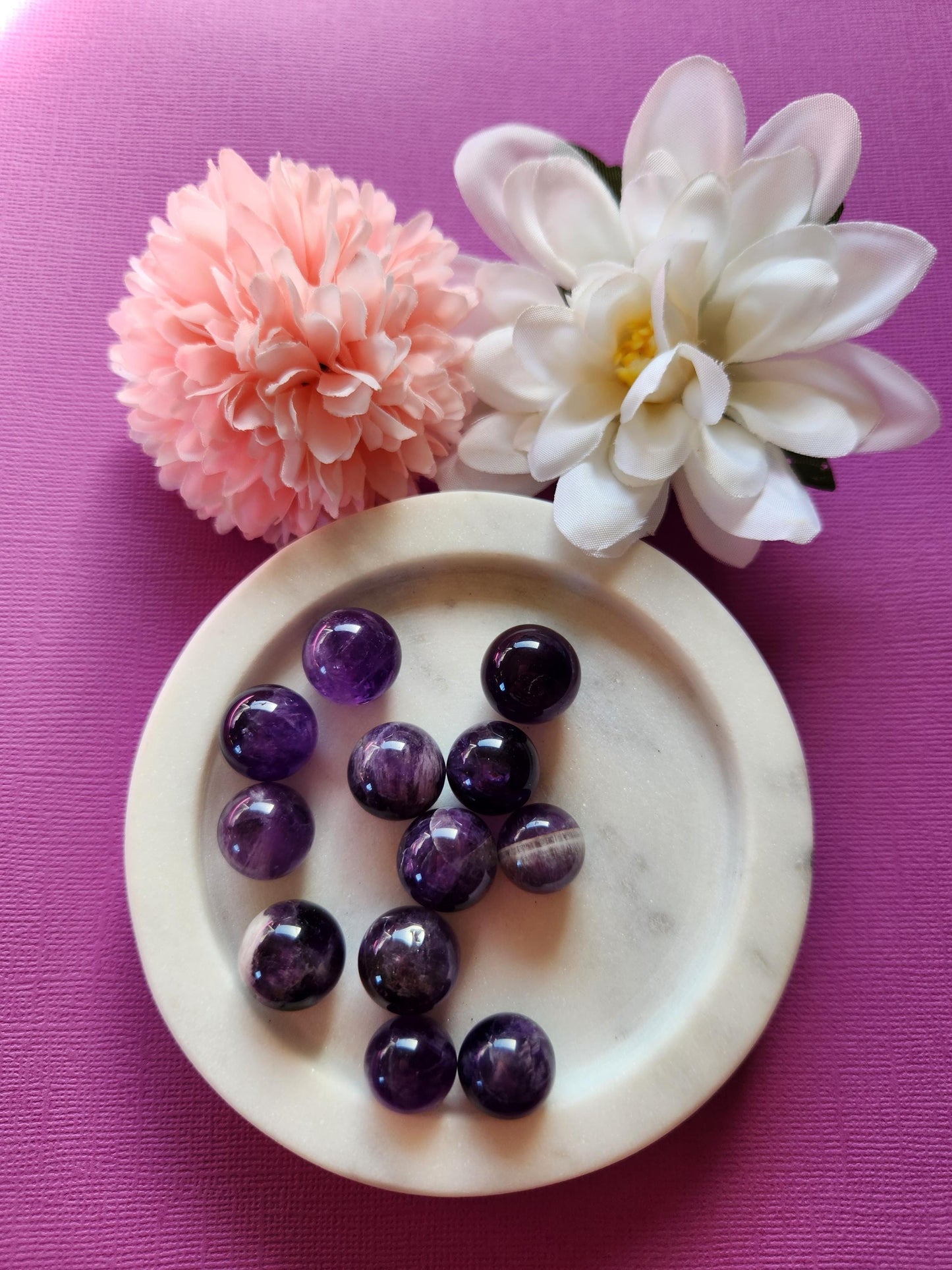 Polished Amethyst mini spheres displayed in a white dish with delicate flowers in the background on a dark purple backdrop, showcasing their rich violet color and smooth finish, great for spiritual work, home decor or gifts