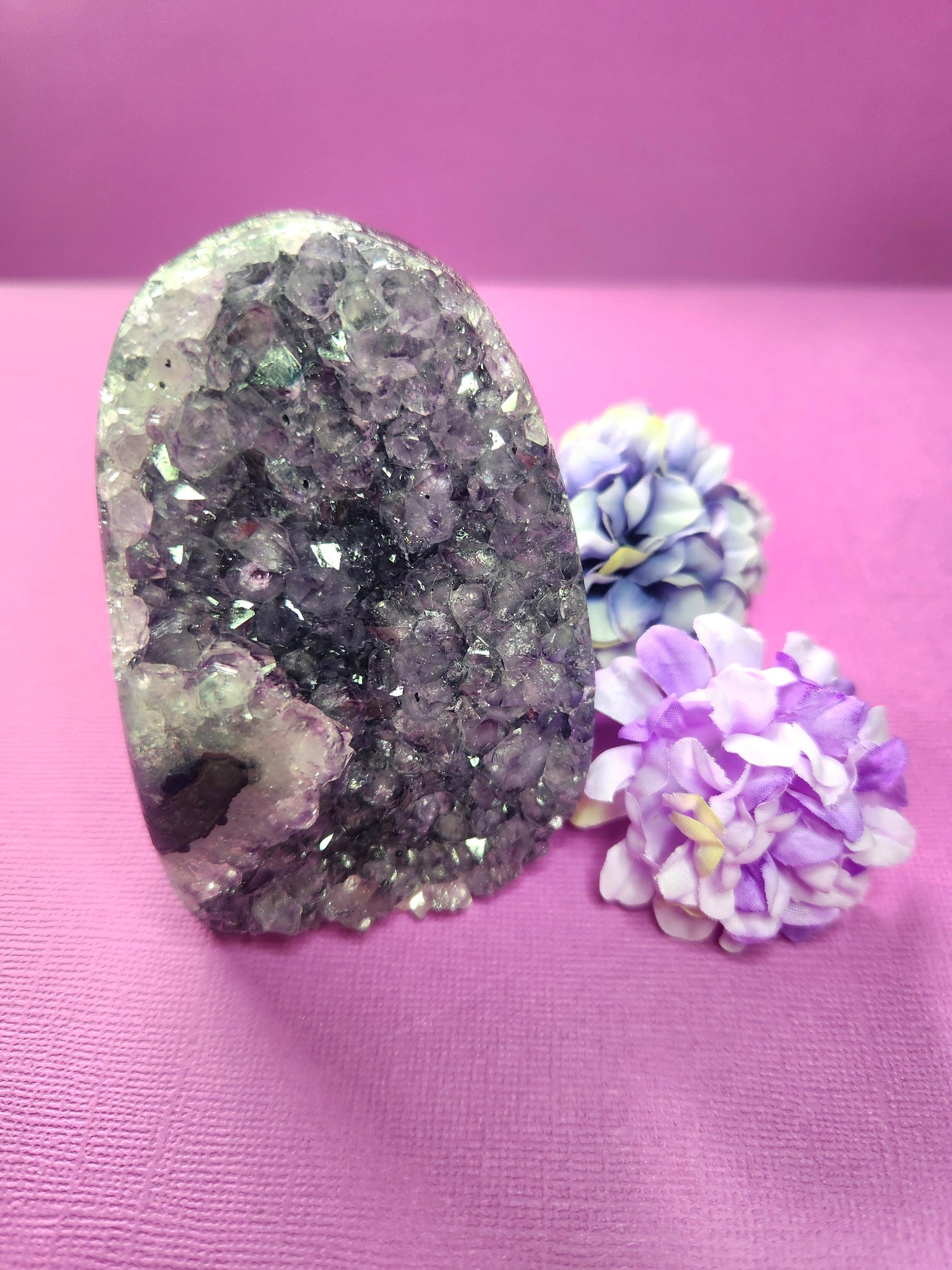 Amethyst Cluster Cut Base with sparkling purple crystals displayed on a purple background with two flower props. Natural raw amethyst geode with flat cut base, perfect for spiritual décor, meditation, and crystal healing. High-quality amethyst crystal for peace, protection, and energy cleansing.