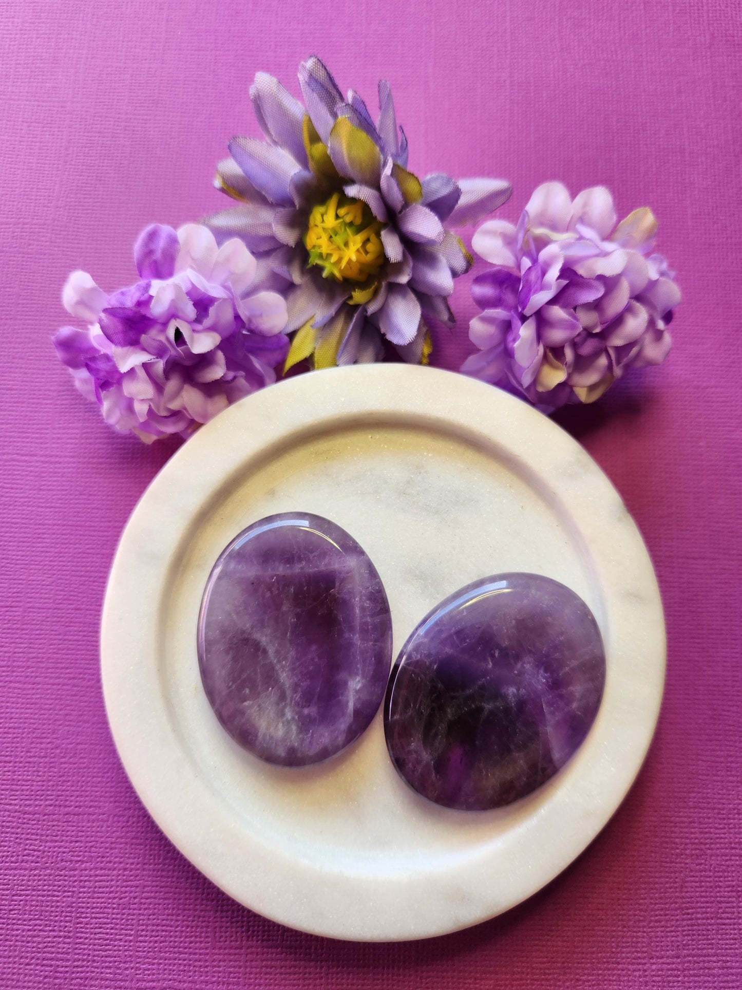Two Amethyst worry stones displayed on a white dish with two flower props, set against a purple background. Calming purple thumb stones for stress relief, intuition, spiritual growth, and crown chakra healing.