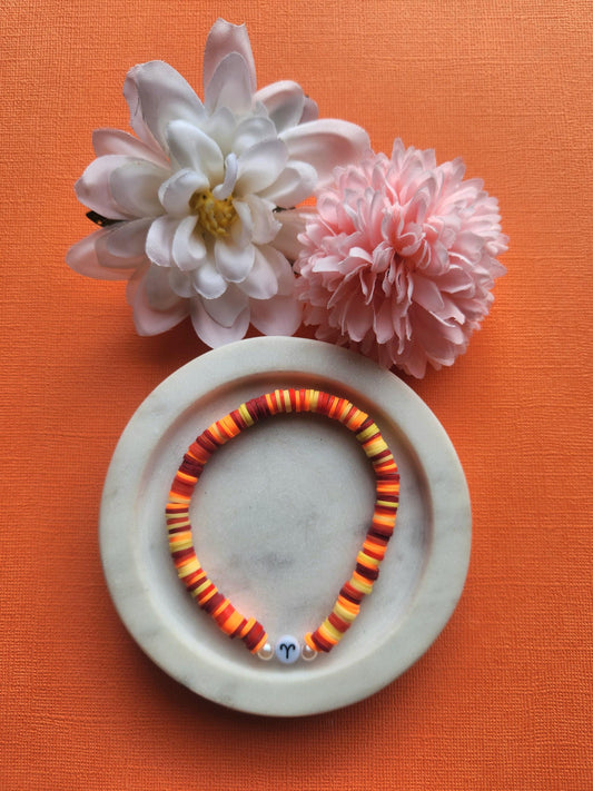 Aries zodiac clay beaded bracelet in white dish with two flower props on orange background, handmade maroon red orange yellow beaded astrology bracelet with Aries symbol bead, stretchy elastic zodiac jewellery product photo