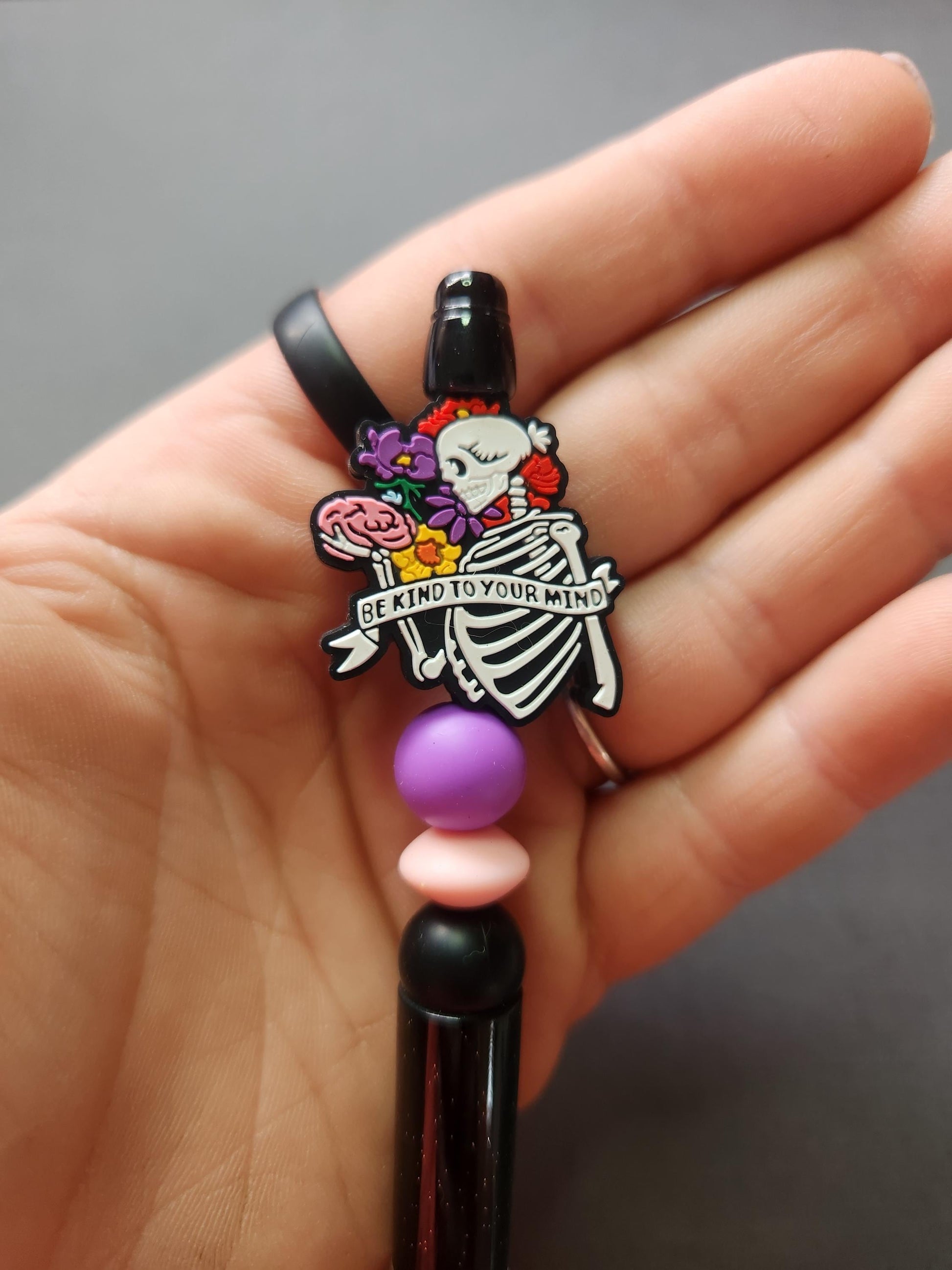 Close-up of Be Kind to Your Mind silicone beaded pen in hand showing skeleton holding brain focal bead with floral banner, pink purple black beads on black glitter pen, detailed product shot on dark grey background, SEO stationery listing image.
