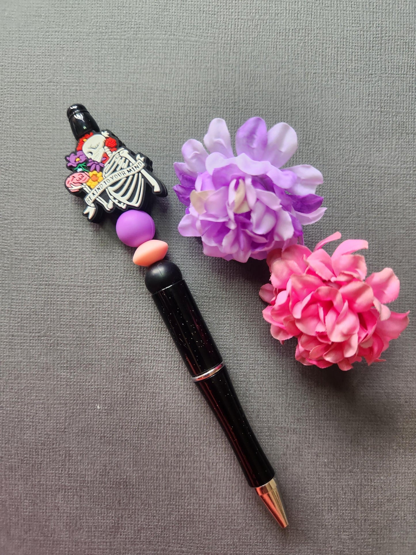 Be Kind to Your Mind silicone beaded pen with skeleton holding brain focal bead, floral banner design, pink purple black silicone beads on black glitter pen, aesthetic stationery photo with flower props on dark grey background, mental health themed beaded pen product image