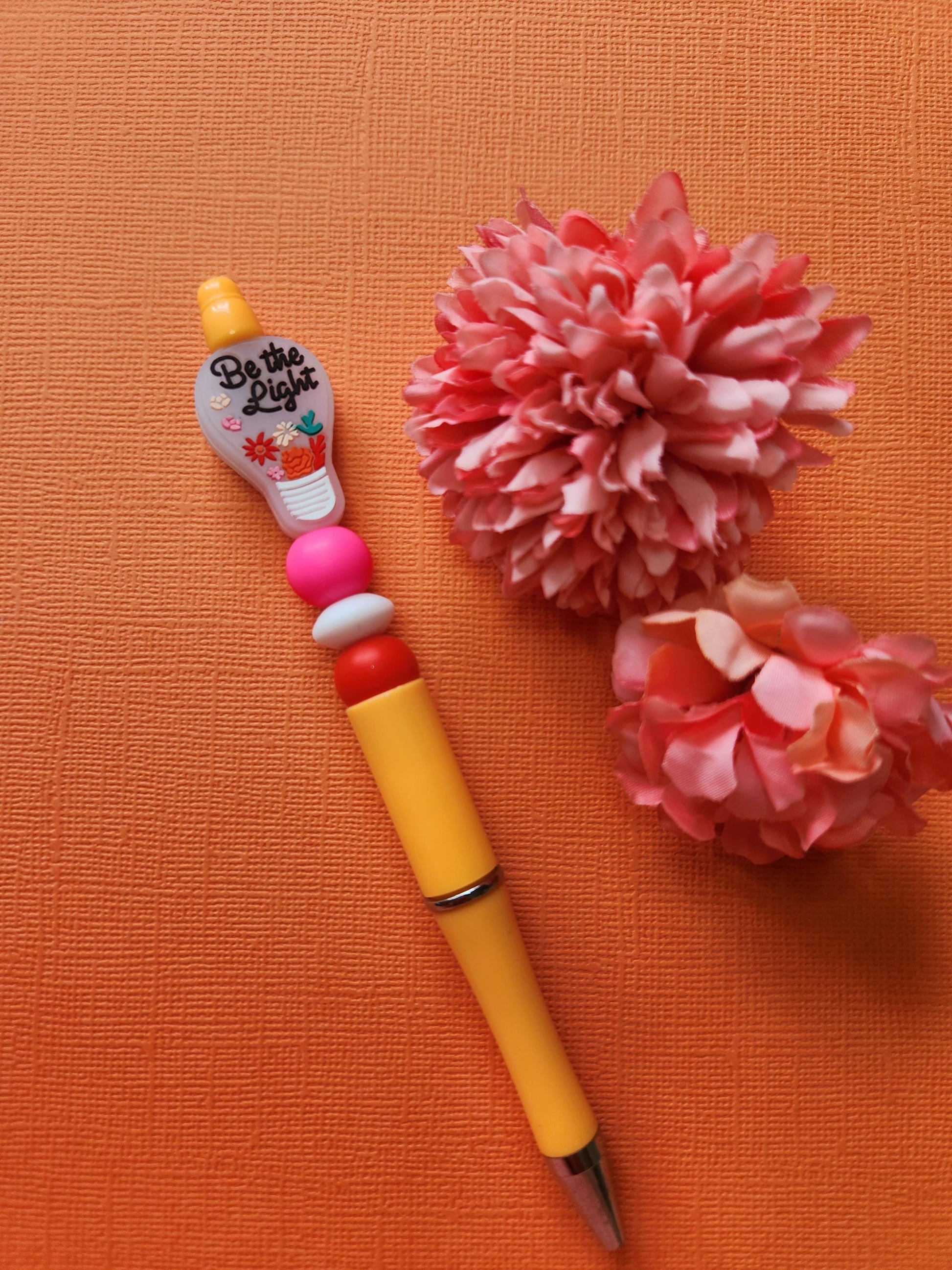 Be the Light silicone beaded pen with light-bulb focal bead featuring floral details and inspirational text, pink white and red silicone beads on an orange pen, styled flat lay with two flower props on an orange background, handmade motivational pen, aesthetic stationery gift