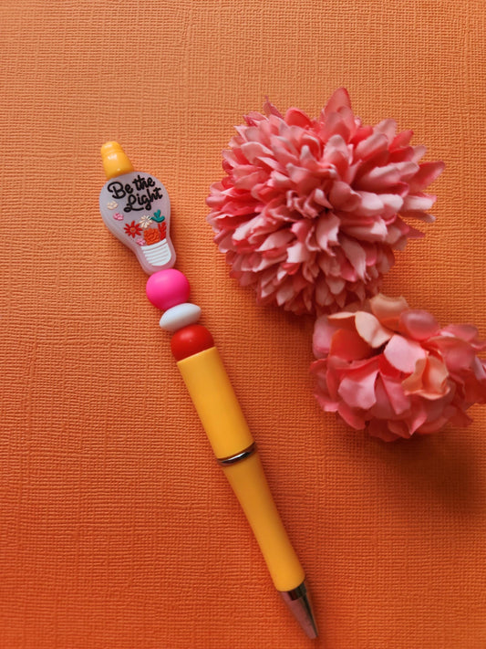 Be the Light silicone beaded pen with light-bulb focal bead featuring floral details and inspirational text, pink white and red silicone beads on an orange pen, styled flat lay with two flower props on an orange background, handmade motivational pen, aesthetic stationery gift