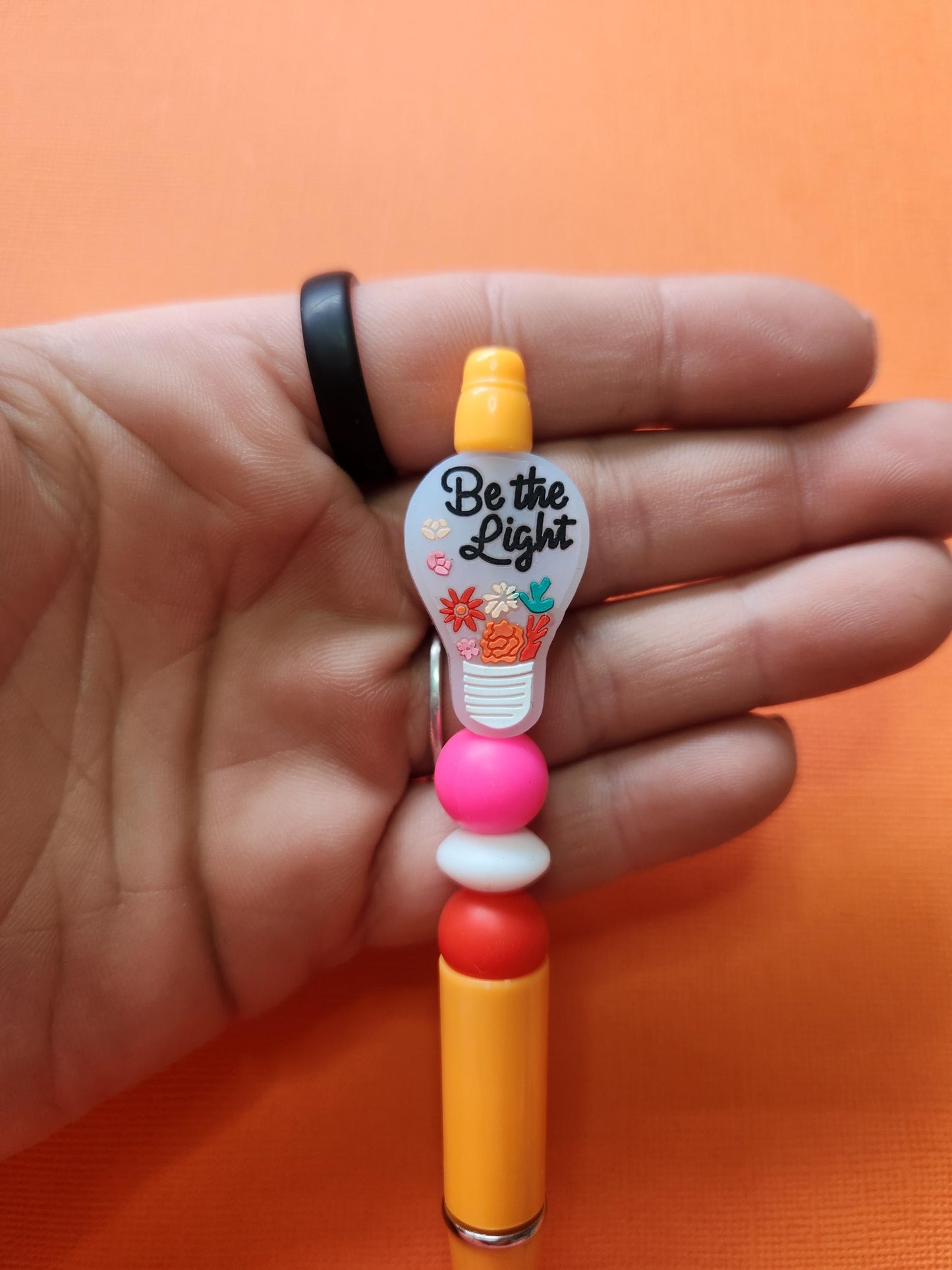 Close-up of Be the Light silicone beaded pen held in hand showing floral light-bulb focal bead with inspirational text, pink white and red silicone beads on orange pen, detailed product photo on orange background, handmade motivational stationery