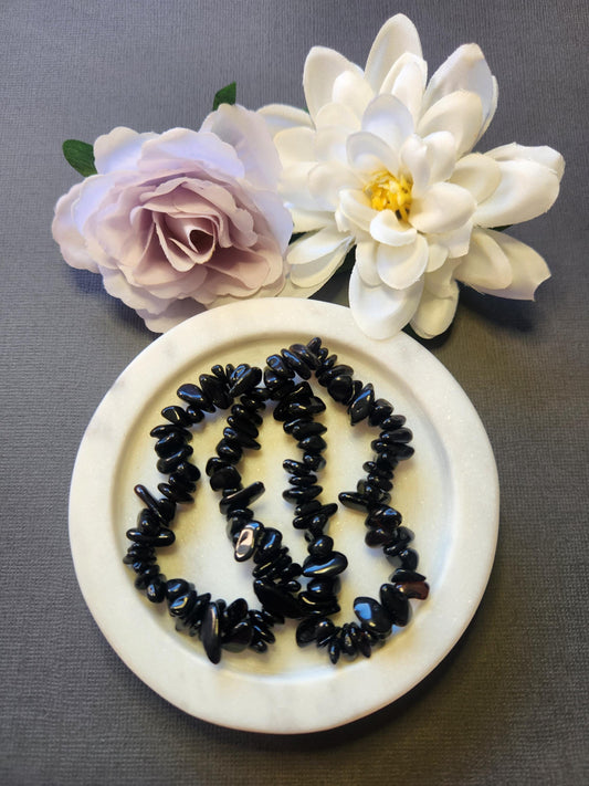 Two Black Obsidian chip bracelets displayed in a white dish with two flower props on a dark grey background, featuring polished black crystal chips that provide grounding, protection, and Root Chakra energy — perfect shielding and healing crystal jewelry gift.