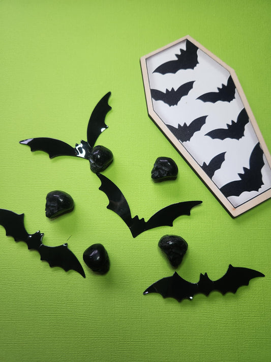 Five polished black obsidian crystal skulls displayed on a bright green backdrop, surrounded by spooky props including black bats and a wooden coffin-shaped decor piece with bat carvings — perfect for Halloween crystal displays, gothic home decor, and metaphysical skull collectors.