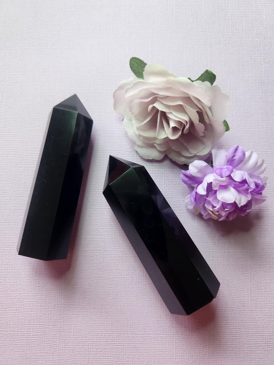 Two polished Black Obsidian crystal towers with glossy deep black finish, standing upright with two flower props on a lavender background, grounding protection crystal points, metaphysical décor, volcanic glass gemstone towers for energy cleansing