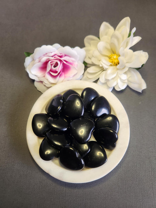 Black obsidian crystal hearts displayed in a white dish on a dark grey background with soft flower props featuring polished black heart stones for protection grounding energy spiritual healing crystal decor and gift