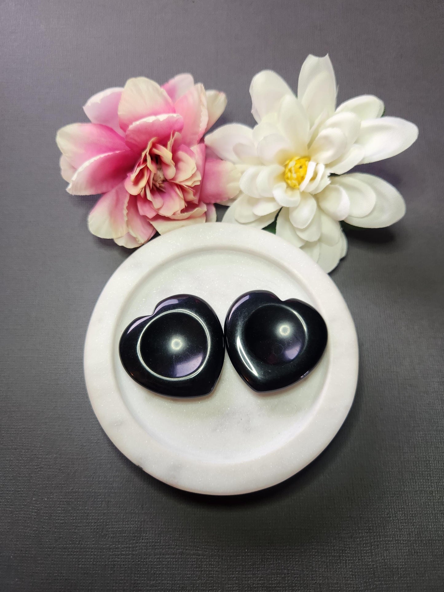 Two black obsidian heart-shaped worry stones displayed in a white dish with two flower props on a dark grey background, polished protection stones for grounding and energy cleansing.