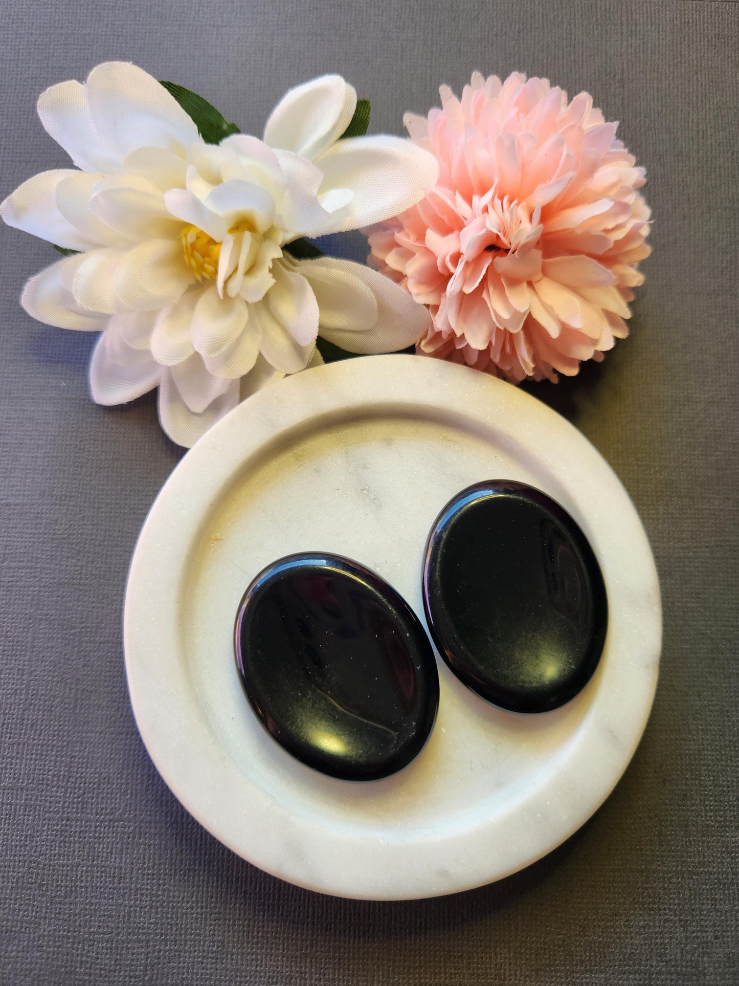 Two polished Black Obsidian worry stones displayed on a white dish with two flower props, set against a grey background. Smooth, glossy black thumb stones used for protection, grounding, and releasing negativity.