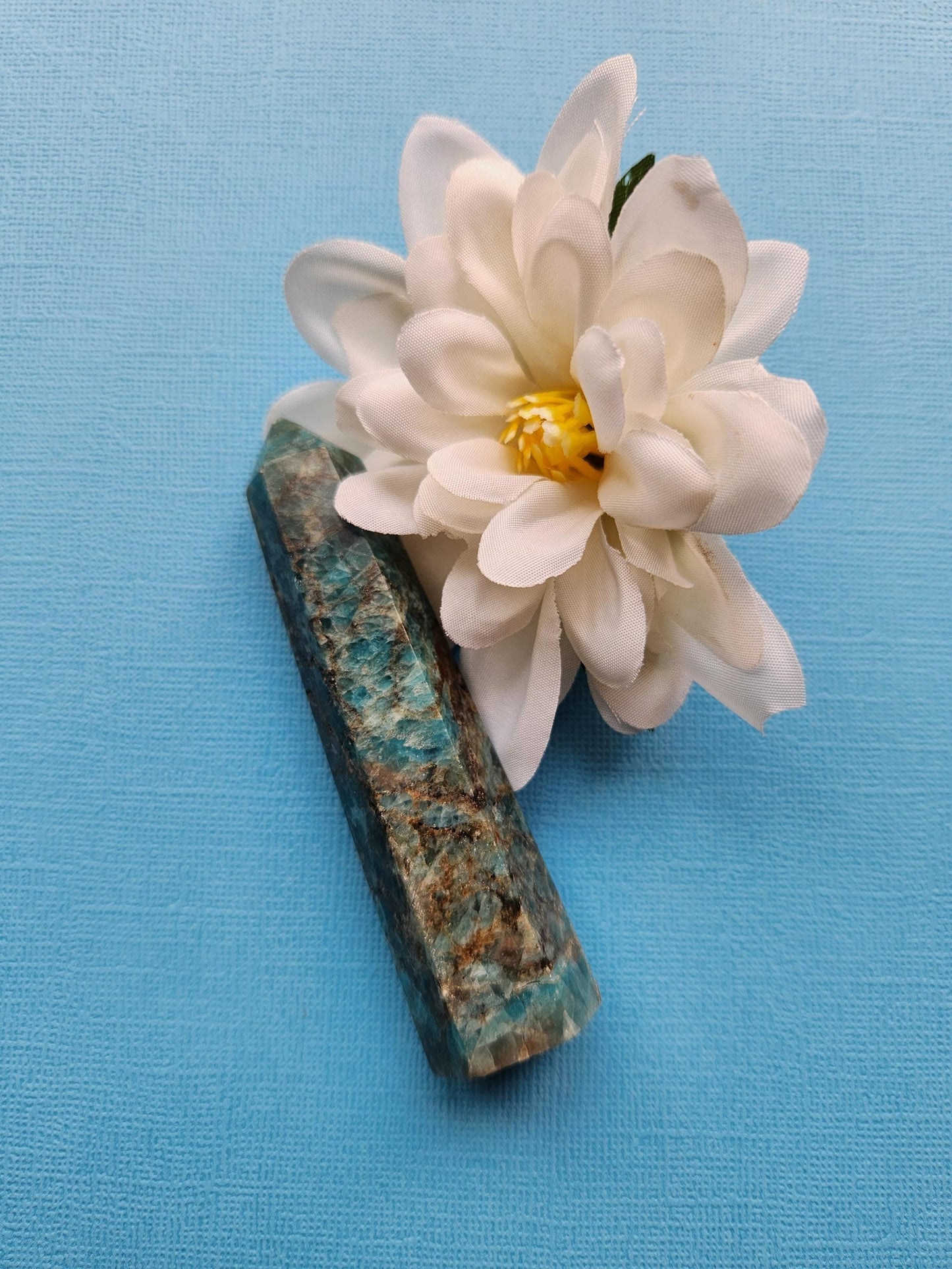 Single Blue Apatite tower photographed at an angle on a blue background, highlighting its polished facets and translucent blue hues. This natural crystal point supports Throat Chakra healing, manifestation, and energy work. The photo showcases the tower’s unique shape and clarity, ideal for meditation spaces, spiritual altars, or crystal collections. The blue background enhances the stone’s vibrant color.