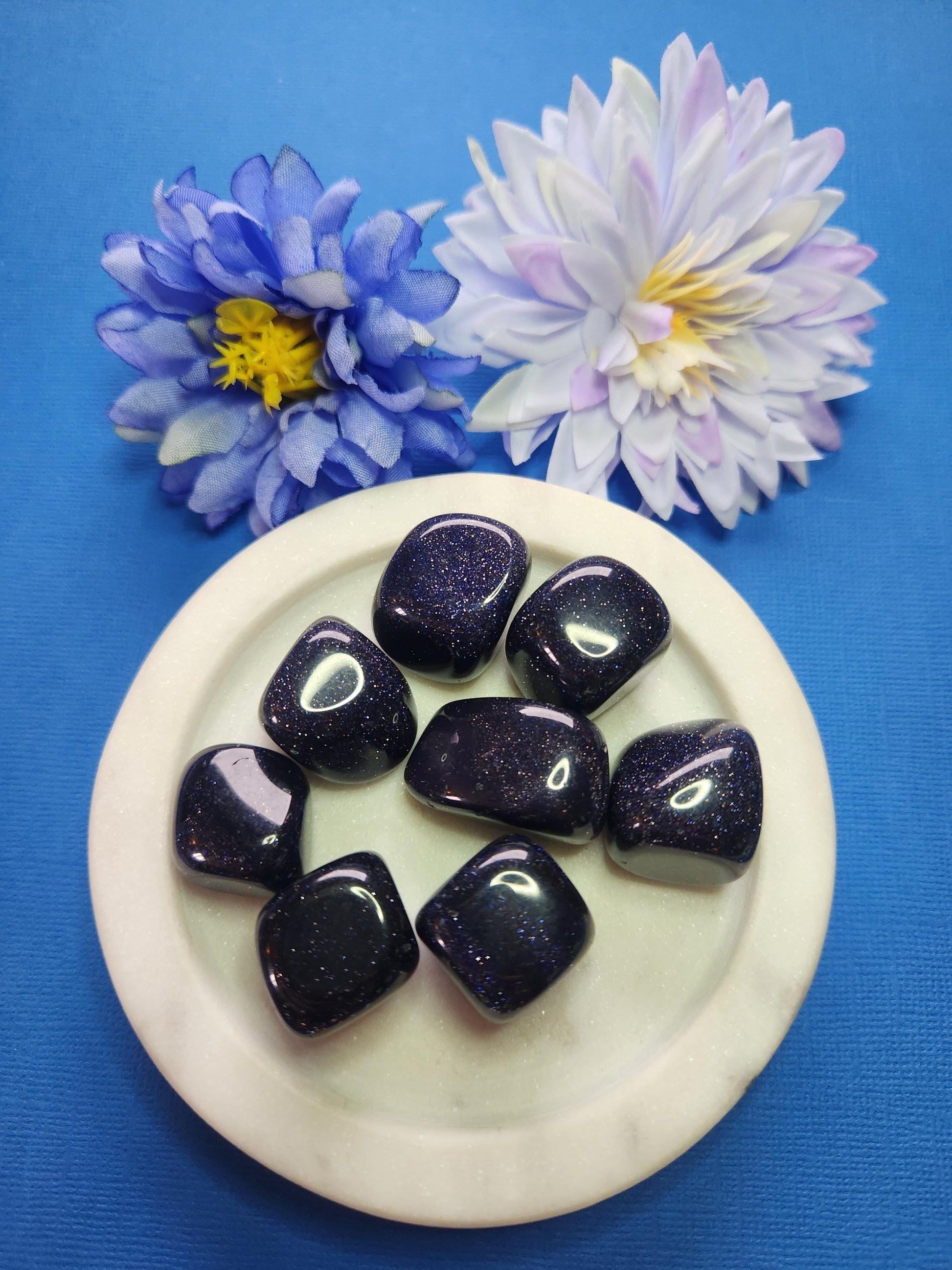 Polished Blue Sandstone crystal tumbles displayed in a white dish on a blue background with two flower props, natural blue gemstone pieces for grounding, calming energy, meditation, chakra balancing, manifestation, spiritual crystal collections, and metaphysical décor
