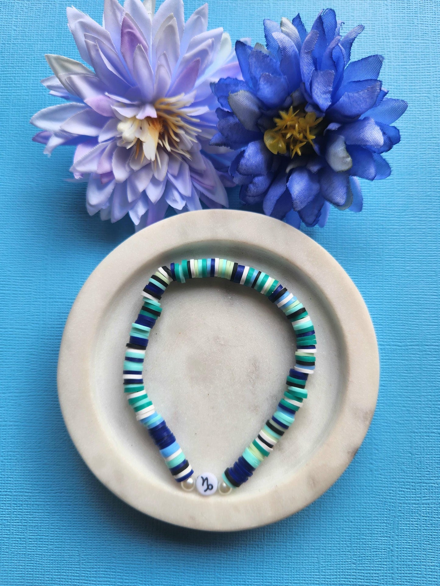 Capricorn zodiac clay beaded bracelet in white dish with two flower props on blue background, handmade navy, blue, white and green polymer clay beads with Capricorn symbol bead, elastic stretch astrology jewellery product photo