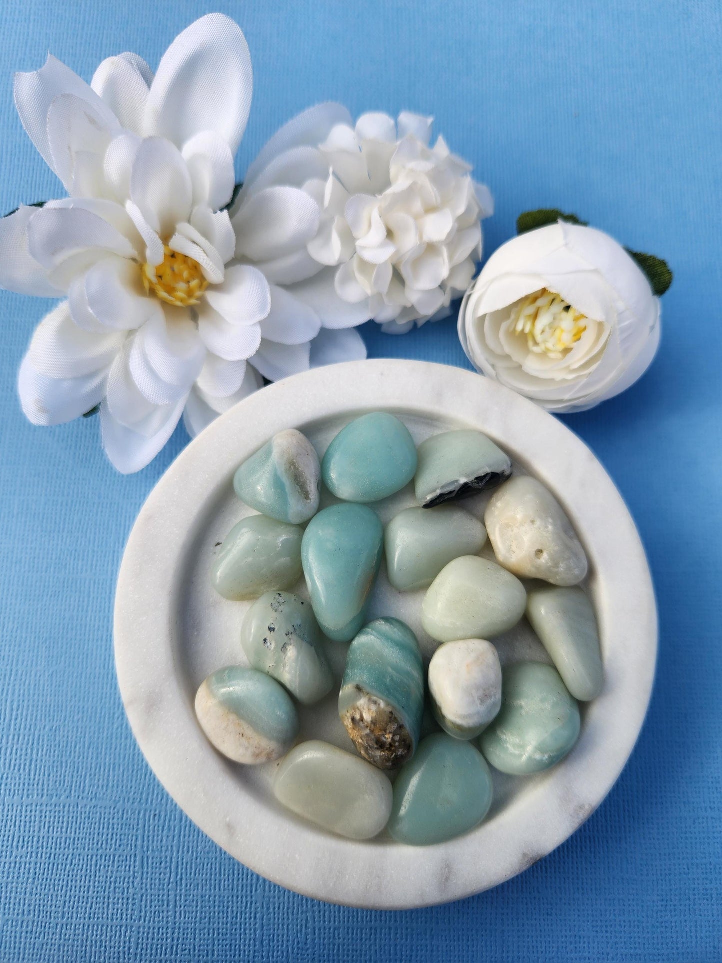 Polished Caribbean Calcite tumble stones in a blue dish, with flower props in the background on a bright blue backdrop, showcasing their calming blue and white swirls.