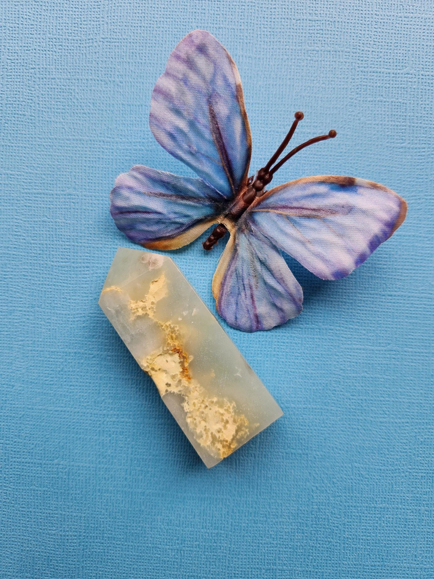 Caribbean Calcite crystal tower displayed on a blue background, showcasing its soft pastel blue and white hues with natural striations and polished facets. This natural crystal point is known for promoting emotional healing, calming energy, and enhancing creativity. Ideal for meditation, spiritual growth, and crystal collections. The blue backdrop highlights the stone’s gentle colors and smooth polished surface.