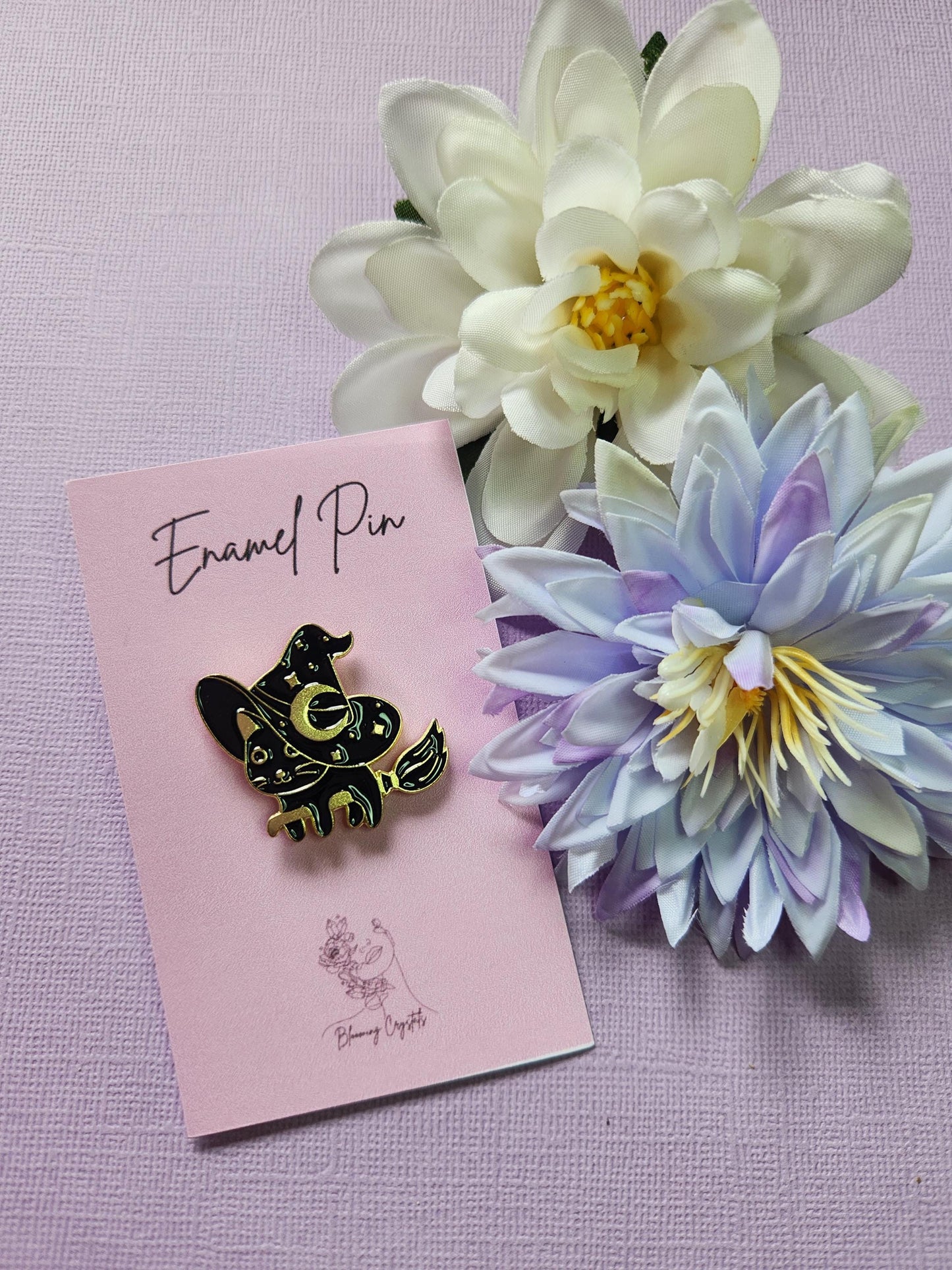 Cute black cat enamel pin with gold outlining, wearing a large witch hat and riding a flying broomstick. Entire pin is black and gold. Styled with two flower props on a lavender background. Witchy cat pin, magical feline enamel pin, spooky cute accessory, Halloween broomstick pin, pastel goth witch pin.