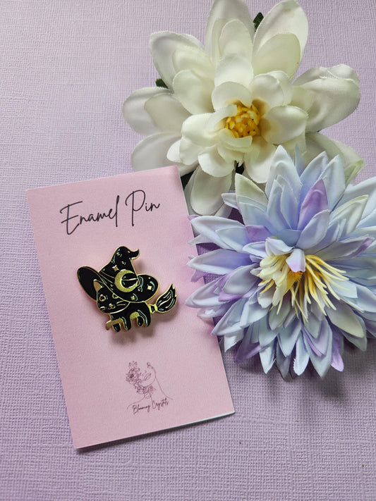 Cute black cat enamel pin with gold outlining, wearing a large witch hat and riding a flying broomstick. Entire pin is black and gold. Styled with two flower props on a lavender background. Witchy cat pin, magical feline enamel pin, spooky cute accessory, Halloween broomstick pin, pastel goth witch pin.