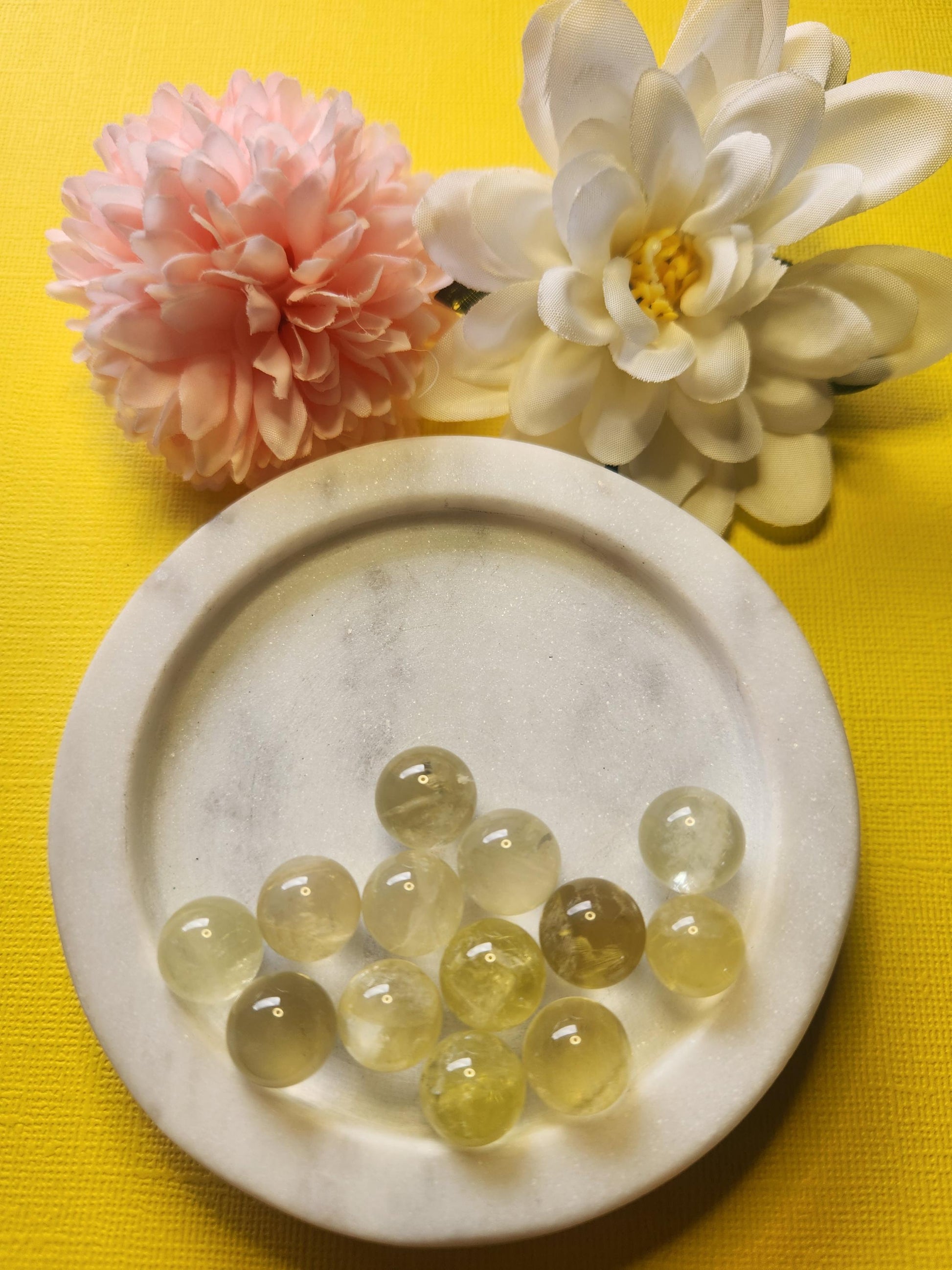 Close-up photo of several 1.2cm citrine mini spheres displayed in a white dish with two soft flower props on a yellow backdrop. The spheres show translucent golden-yellow hues with a polished glossy finish. Ideal for citrine crystals, mini crystal spheres, crystal collections, and decorative gemstone accents