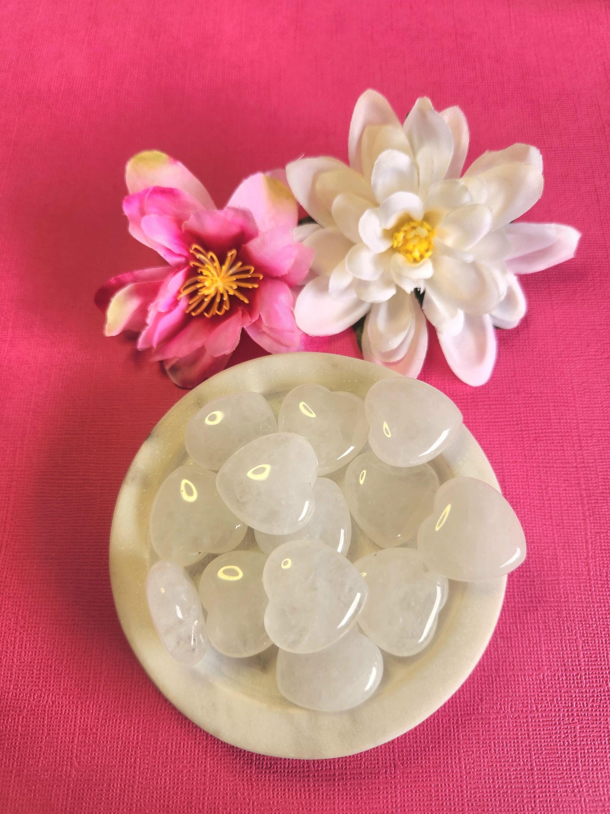 Clear quartz crystal hearts displayed in a white dish on a pink background with soft flower props featuring polished heart stones for healing energy clarity spiritual balance crystal decor and gift