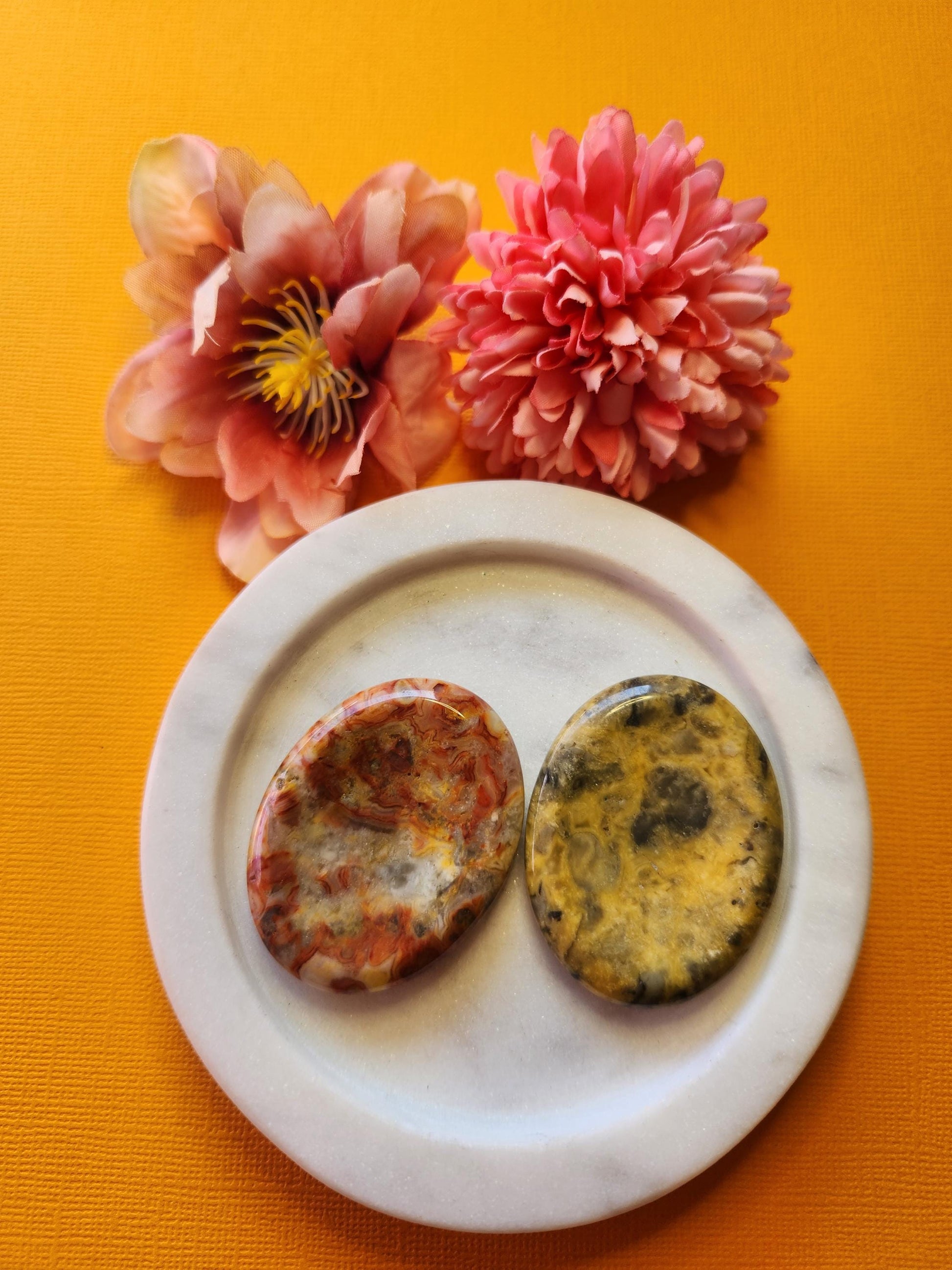Two Crazy Lace Agate worry stones displayed on a white dish with two flower props, set against a yellow background. Polished thumb stones for joy, grounding, emotional balance, and positive energy.
