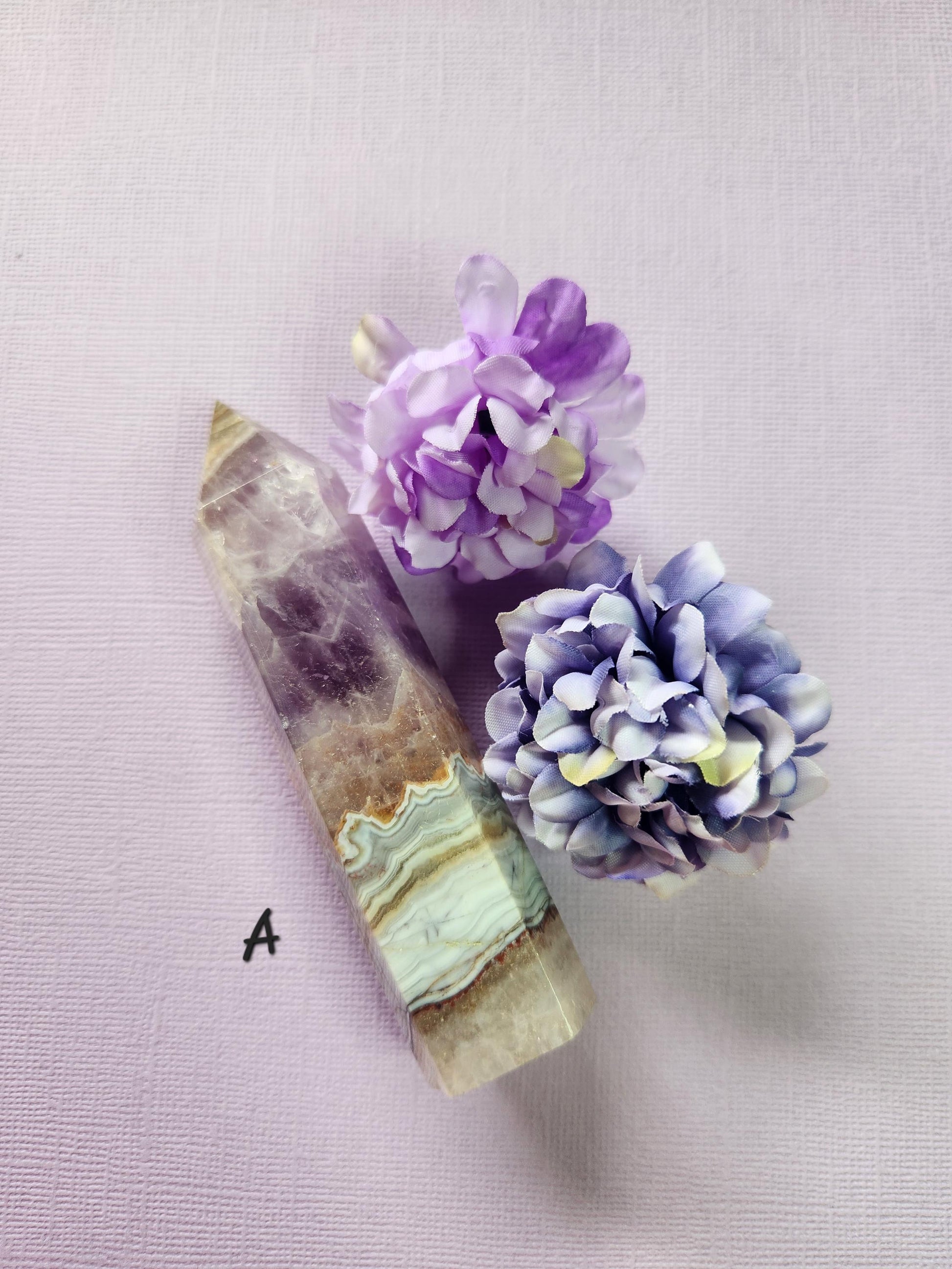 Close-up of Tower A: Amethyst and Crazy Lace Agate Crystal Tower on a lavender background with two flower props. Polished natural gemstone tower featuring deep purple amethyst and orange crazy lace agate patterns. Healing crystal for meditation, chakra balancing, energy cleansing, spiritual décor, and crystal collections.