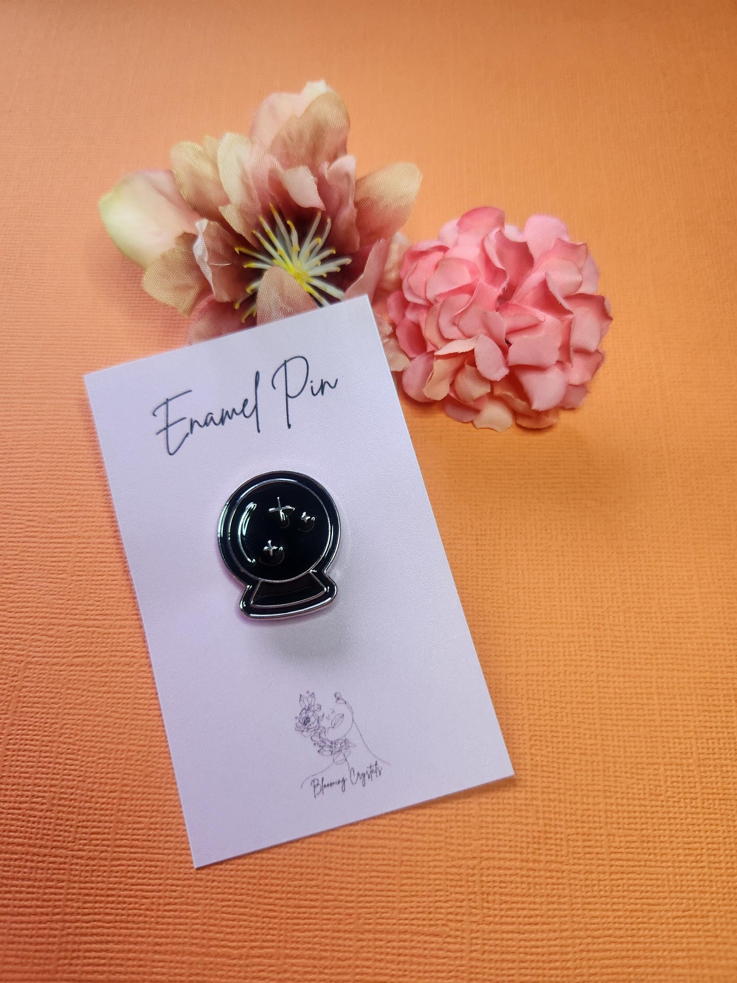 Black crystal ball enamel pin with silver outline and silver star accents, styled with floral props on a bright orange background. Witchy aesthetic pin, mystical accessories, celestial enamel pin, crystal ball witch pin, Halloween jewelry flat lay.