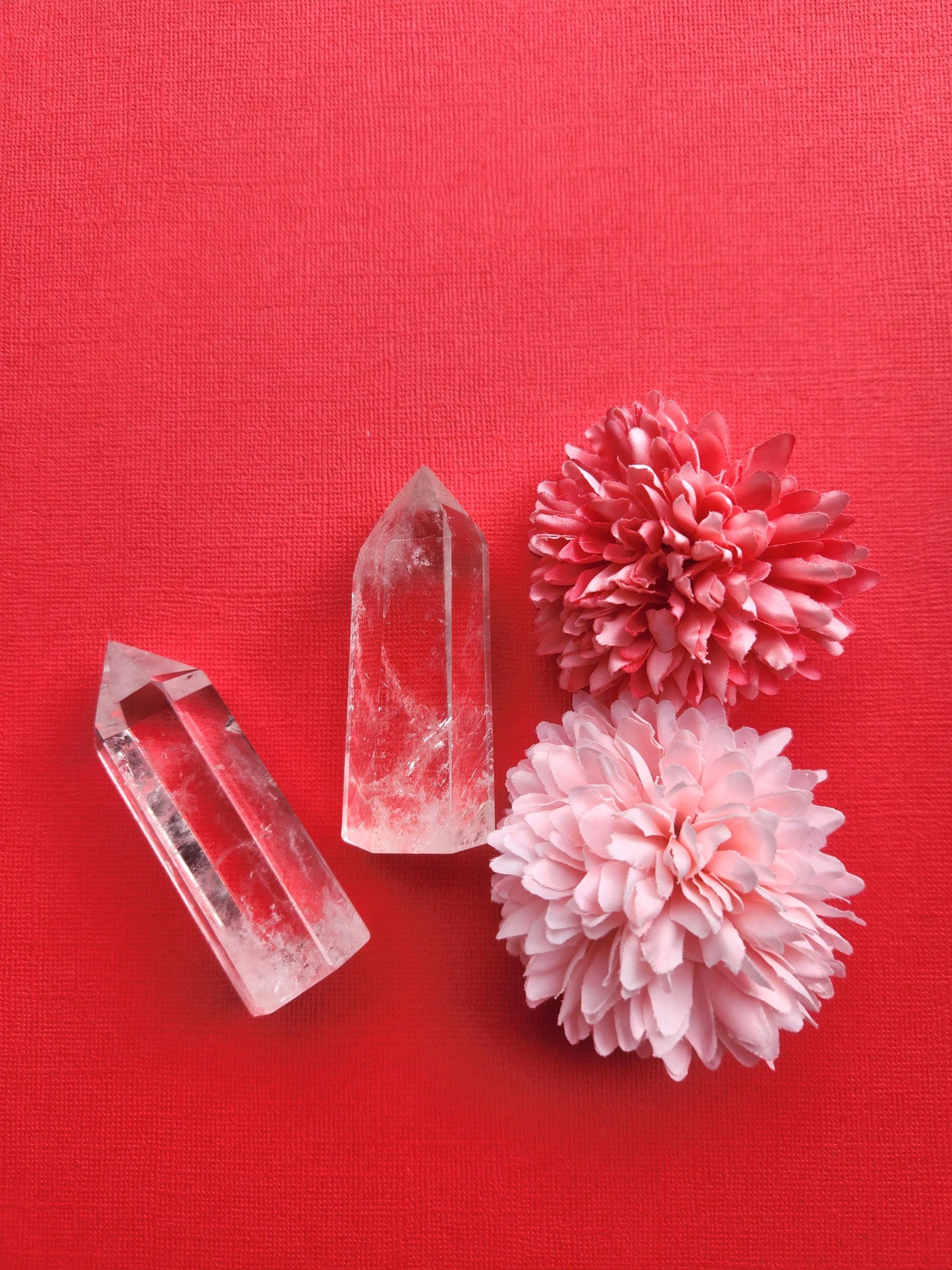Two polished Clear Quartz crystal towers displayed together on a red background with two flower props, natural clear quartz points for energy amplification, healing, meditation, spiritual growth, chakra balancing, manifestation, Reiki, home altar décor, metaphysical crystal collections, and crystal healing practices