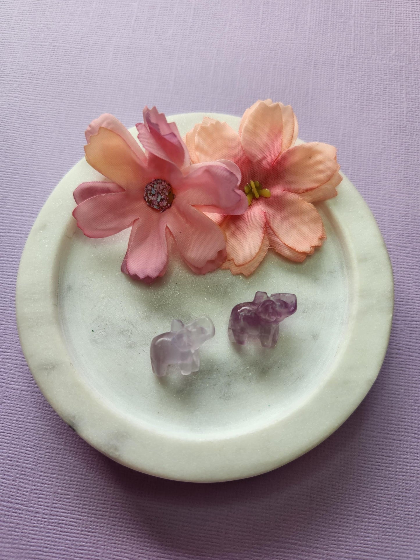 Close-up photo of two fluorite mini elephant carvings displayed in a white dish with two soft flower props on a lavender backdrop. The elephants feature natural purple, green and blue fluorite banding with a polished glossy finish. Perfect for fluorite carvings, mini crystal animals, elephant crystal decor and collectible gemstone figures.