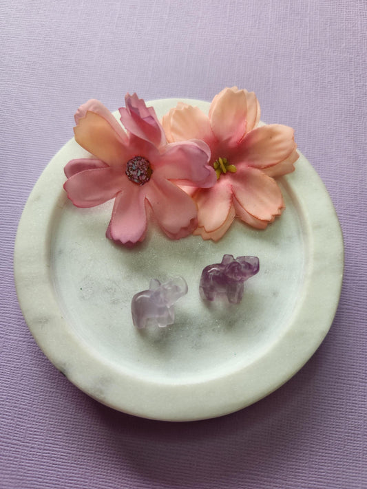 Close-up photo of two fluorite mini elephant carvings displayed in a white dish with two soft flower props on a lavender backdrop. The elephants feature natural purple, green and blue fluorite banding with a polished glossy finish. Perfect for fluorite carvings, mini crystal animals, elephant crystal decor and collectible gemstone figures.