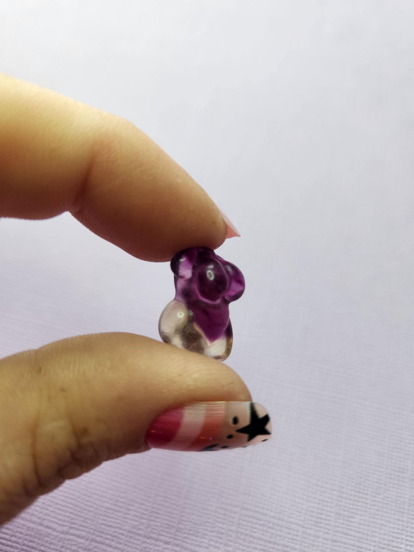Close-up photo of a single fluorite mini goddess body carving held between fingers, showing smooth polished details and natural purple, green and blue fluorite banding against a lavender backdrop. Perfect for fluorite carvings, goddess body crystals, collectible gemstone figures and pocket-sized crystal pieces.
