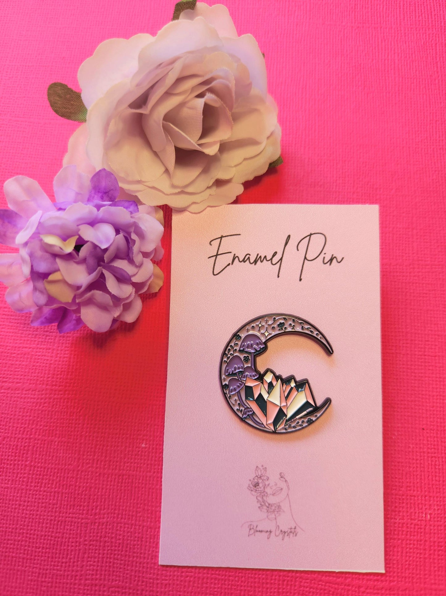 Crystal Moon enamel pin featuring a purple crescent moon with purple mushrooms and pink, white, and dark blue crystals, displayed on a pink backing card with flower props and a bright pink backdrop.