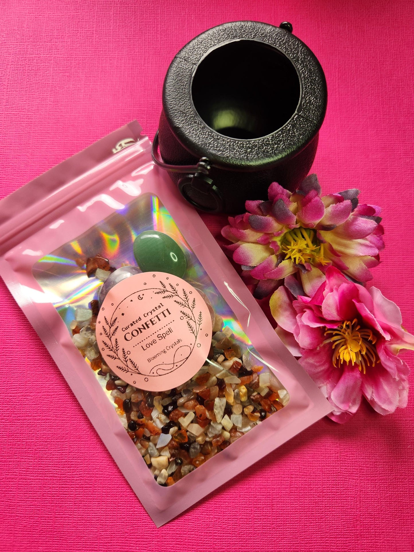 Love spell crystal confetti packaged in a pink holographic bag with a label. Includes crystal chips—Carnelian, Peach Moonstone, Garnet—and tumbled stones of Rose Quartz, Green Aventurine, and Amethyst. Comes with a black cauldron and is styled with two flower props on a bright pink background. Witchy love spell kit, self-love crystal mix, attraction ritual supplies, magical crystal confetti gift.