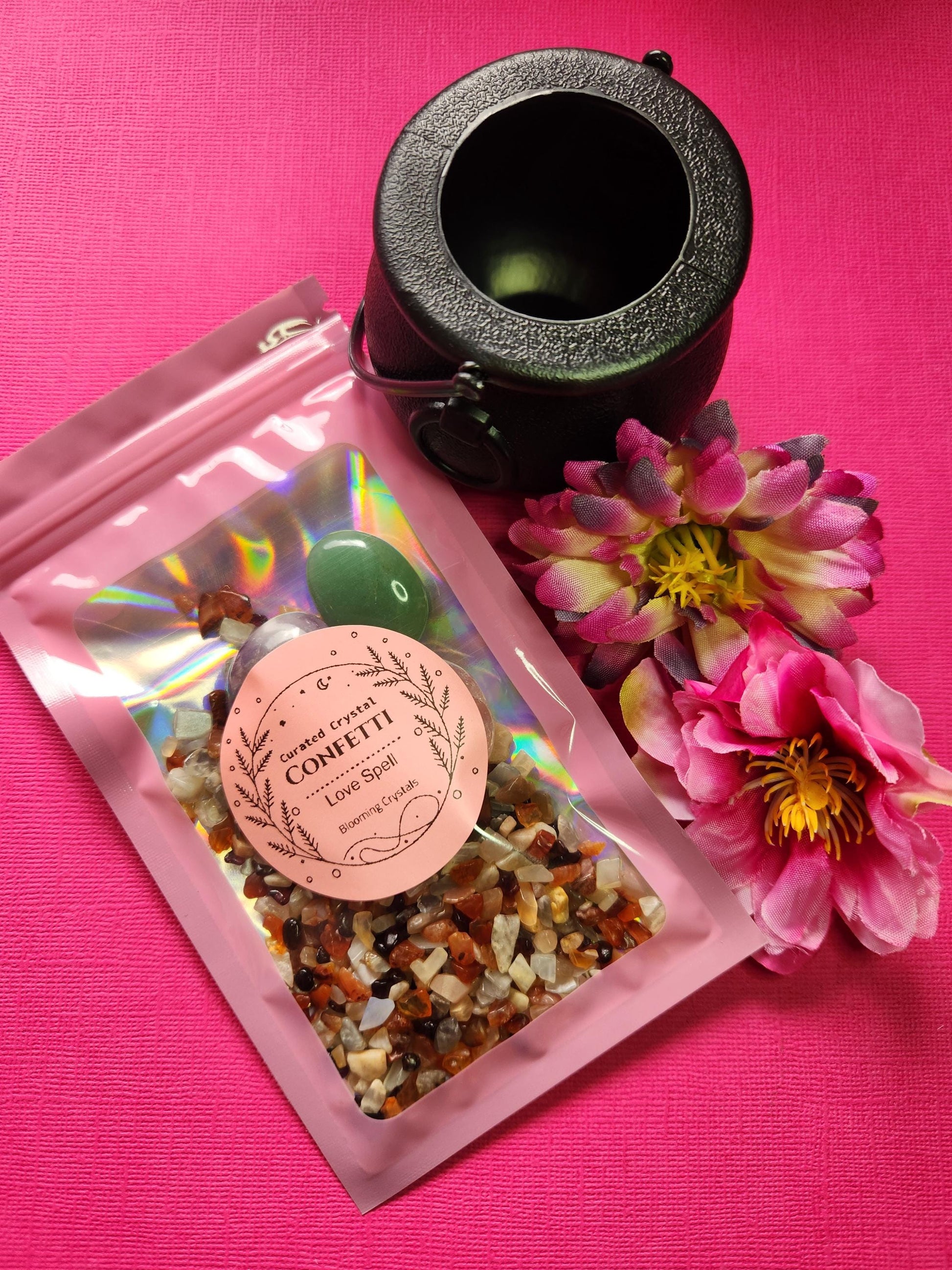 Love spell crystal confetti packaged in a pink holographic bag with a label. Includes crystal chips—Carnelian, Peach Moonstone, Garnet—and tumbled stones of Rose Quartz, Green Aventurine, and Amethyst. Comes with a black cauldron and is styled with two flower props on a bright pink background. Witchy love spell kit, self-love crystal mix, attraction ritual supplies, magical crystal confetti gift.