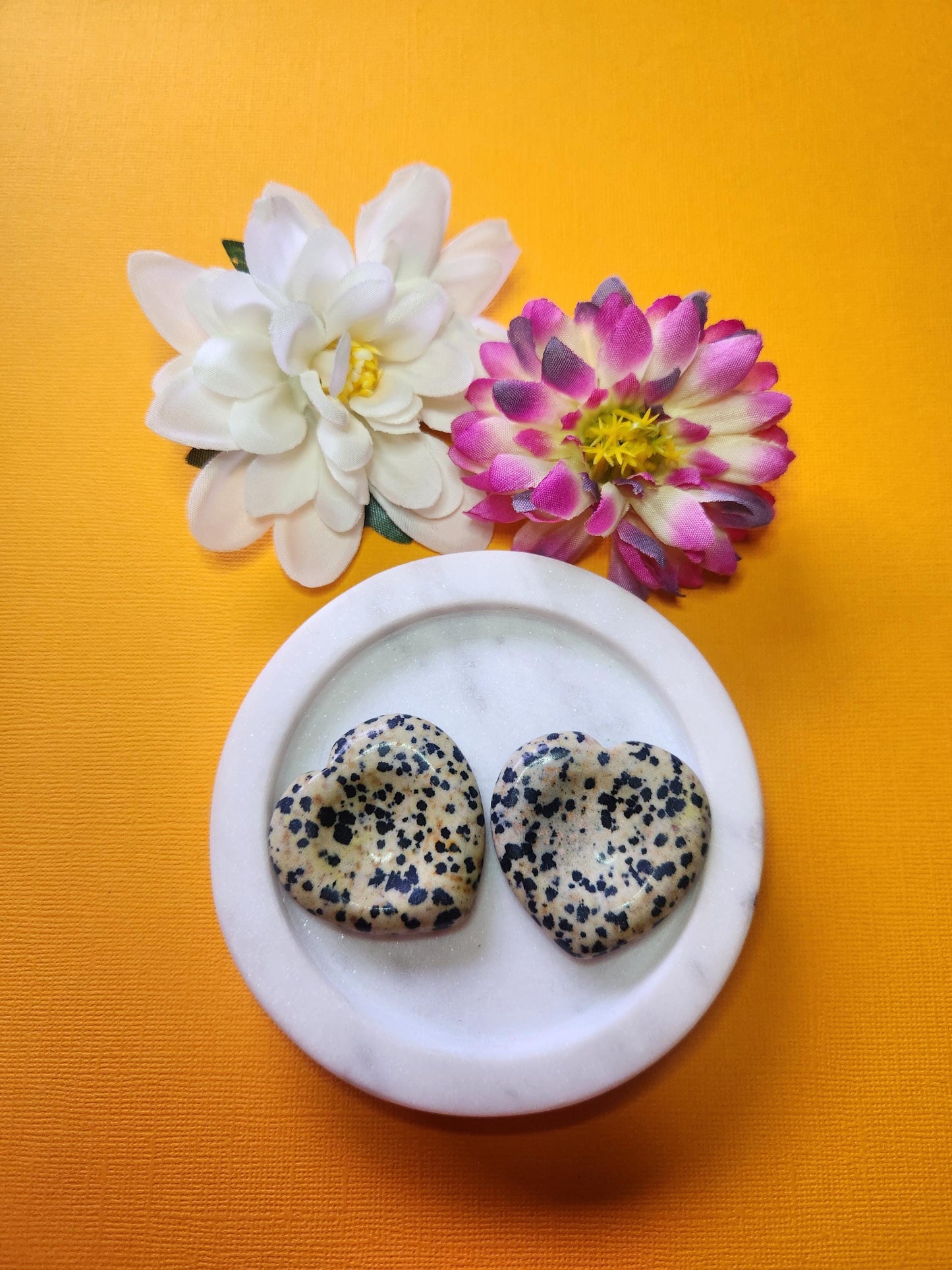 Two Dalmatian Jasper heart-shaped worry stones displayed in a white dish with two floral props on a soft yellow background, grounding crystal hearts for stress relief and mindfulness.