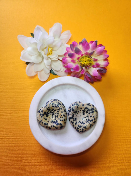 Two Dalmatian Jasper heart-shaped worry stones displayed in a white dish with two floral props on a soft yellow background, grounding crystal hearts for stress relief and mindfulness.