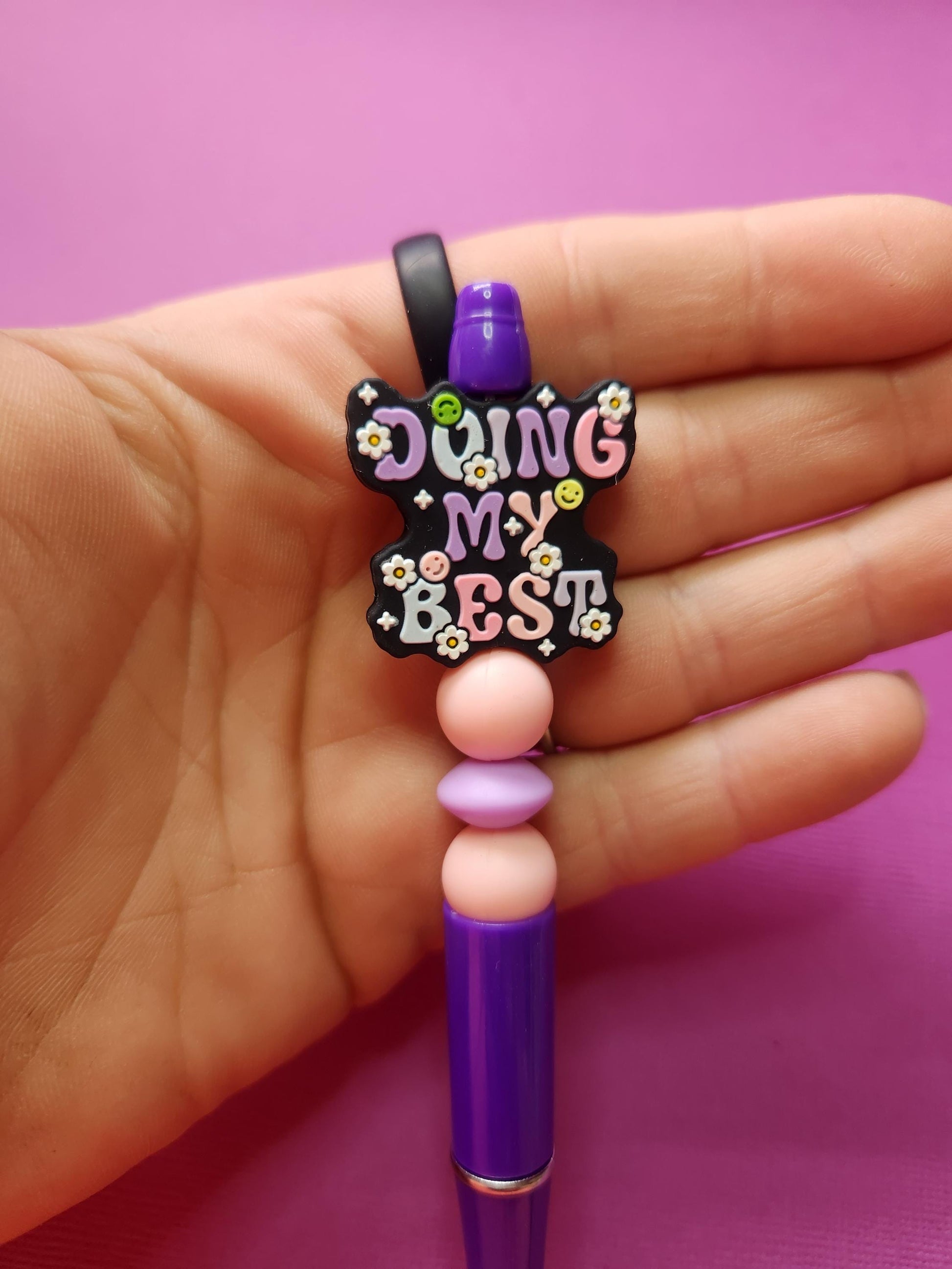Close-up of the Doing My Best silicone beaded pen held in hand, showing the black focal bead with pastel lavender, baby blue and light pink lettering, with light pink beads and lavender spacer on a purple pen against a purple backdrop. SEO: inspirational pen, silicone bead pen detail, handmade stationery shop, aesthetic writing tools, pastel office accessories, motivational study supplies, Australian small business pen