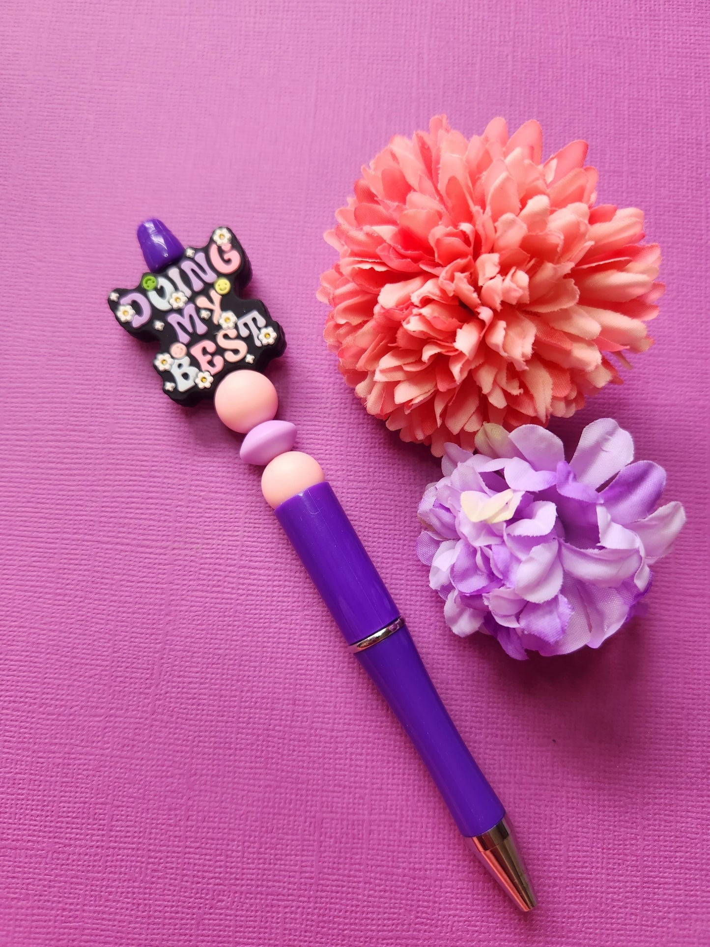 Doing My Best silicone beaded pen with a black focal bead featuring pastel lavender, baby blue and light pink lettering, paired with two light pink beads and a lavender spacer bead on a purple twist-to-write pen, styled with two flower props on a purple background. SEO: motivational pen, pastel aesthetic stationery, silicone beaded pen, handmade pen Australia, cute writing pen, kawaii office supplies, gift for students, desk accessories