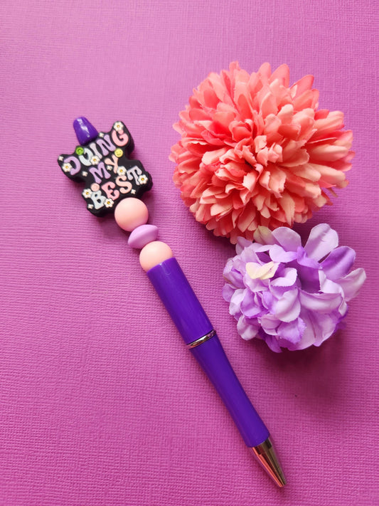 Doing My Best silicone beaded pen with a black focal bead featuring pastel lavender, baby blue and light pink lettering, paired with two light pink beads and a lavender spacer bead on a purple twist-to-write pen, styled with two flower props on a purple background. SEO: motivational pen, pastel aesthetic stationery, silicone beaded pen, handmade pen Australia, cute writing pen, kawaii office supplies, gift for students, desk accessories