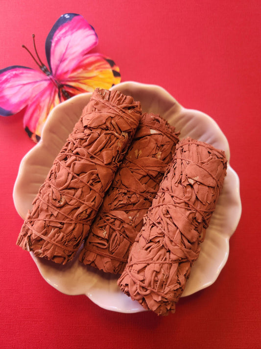 Three Dragon’s Blood Sage smudge sticks displayed in a pink flower dish with a pink and yellow butterfly as props on a red background, showcasing the rich red resin and dried sage for cleansing, energy clearing, and spiritual rituals.