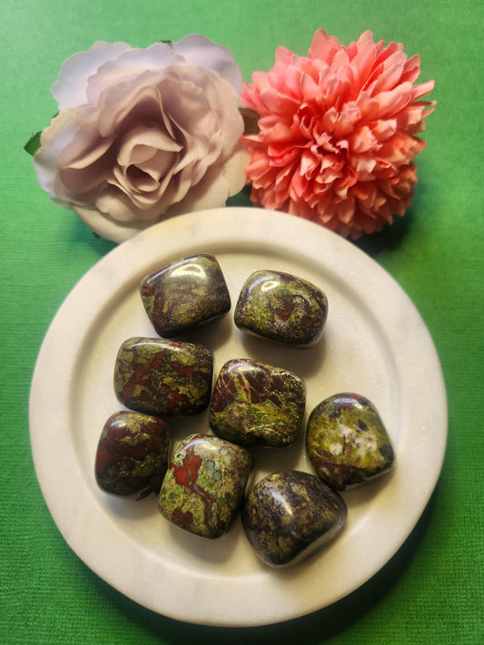 Close-up photo of several Dragons Bloodstone tumbles displayed in a white dish with two soft flower props on a green backdrop. The tumbles show natural dark green and red mottled patterns with a polished glossy finish. Ideal for Dragons Bloodstone crystals, tumbled gemstones, crystal collections, and decorative crystal accents