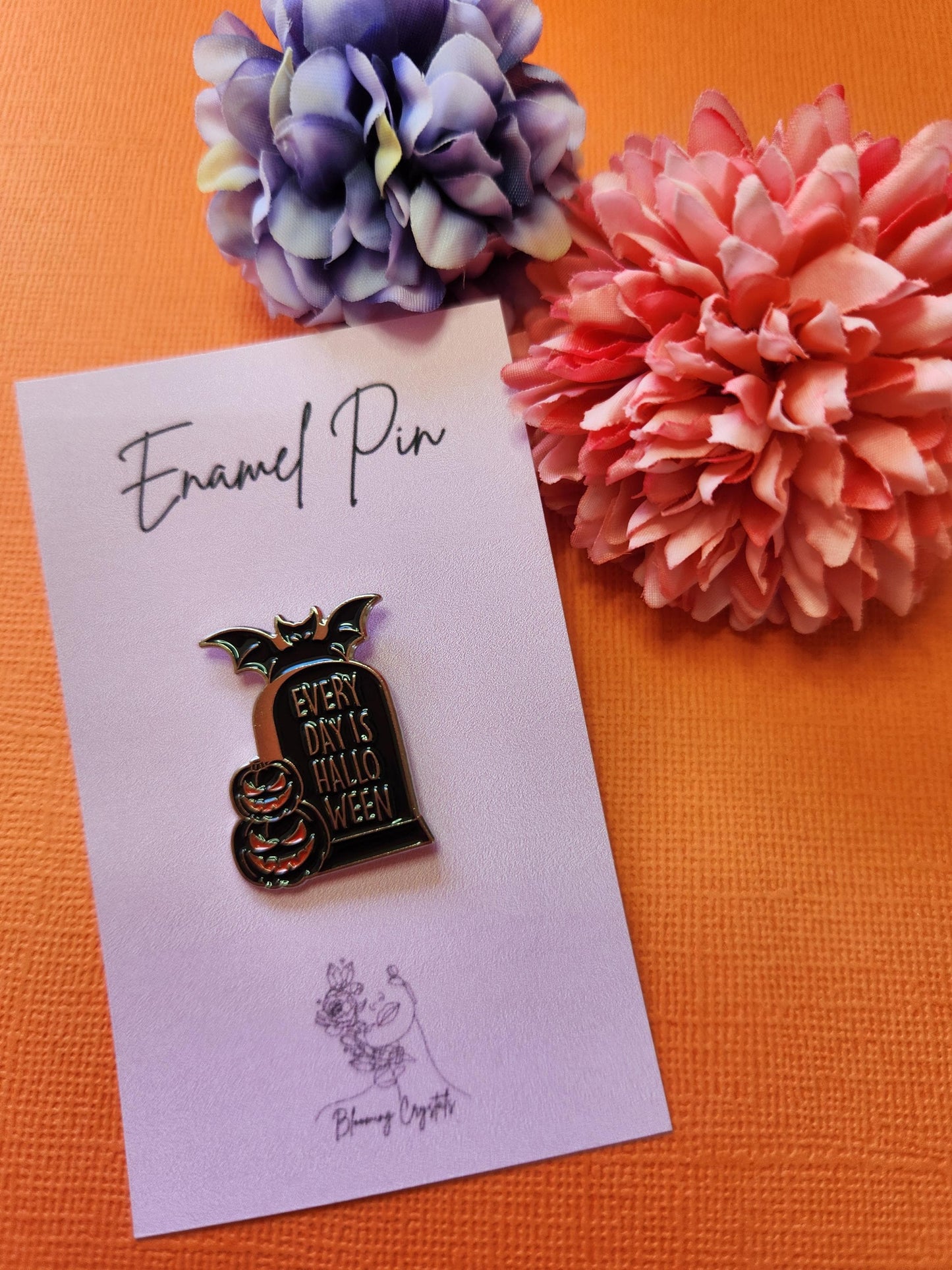 Black enamel pin shaped like a tombstone with silver text that reads "Everyday is Halloween." Features two pumpkins at the base and a bat perched on top. Styled with two flower props on a bright orange background. Halloween enamel pin, spooky tombstone pin, gothic accessory, witchy fall decor, pumpkin and bat pin for Halloween lovers.
