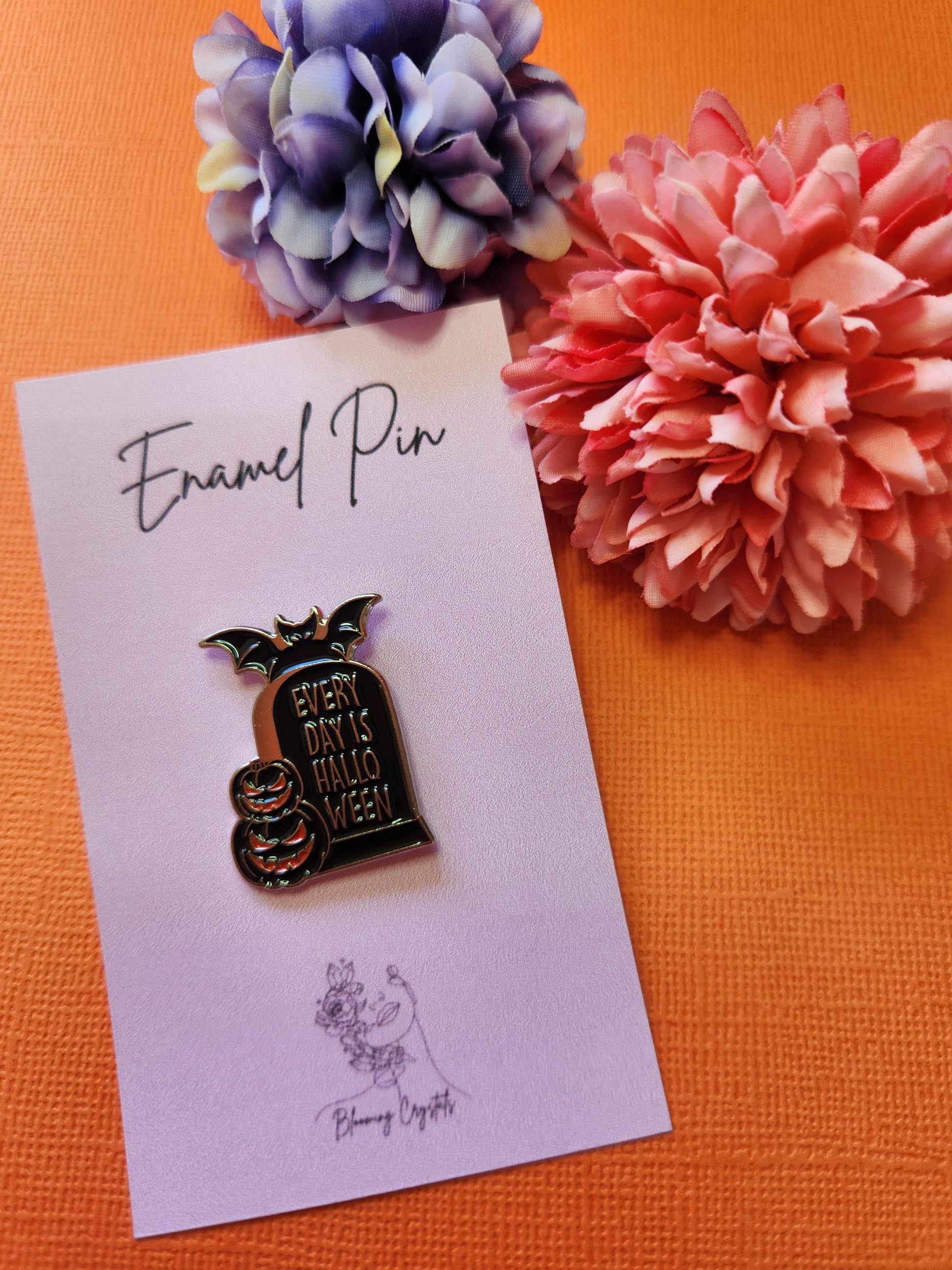 Black enamel pin shaped like a tombstone with silver text that reads "Everyday is Halloween." Features two pumpkins at the base and a bat perched on top. Styled with two flower props on a bright orange background. Halloween enamel pin, spooky tombstone pin, gothic accessory, witchy fall decor, pumpkin and bat pin for Halloween lovers.
