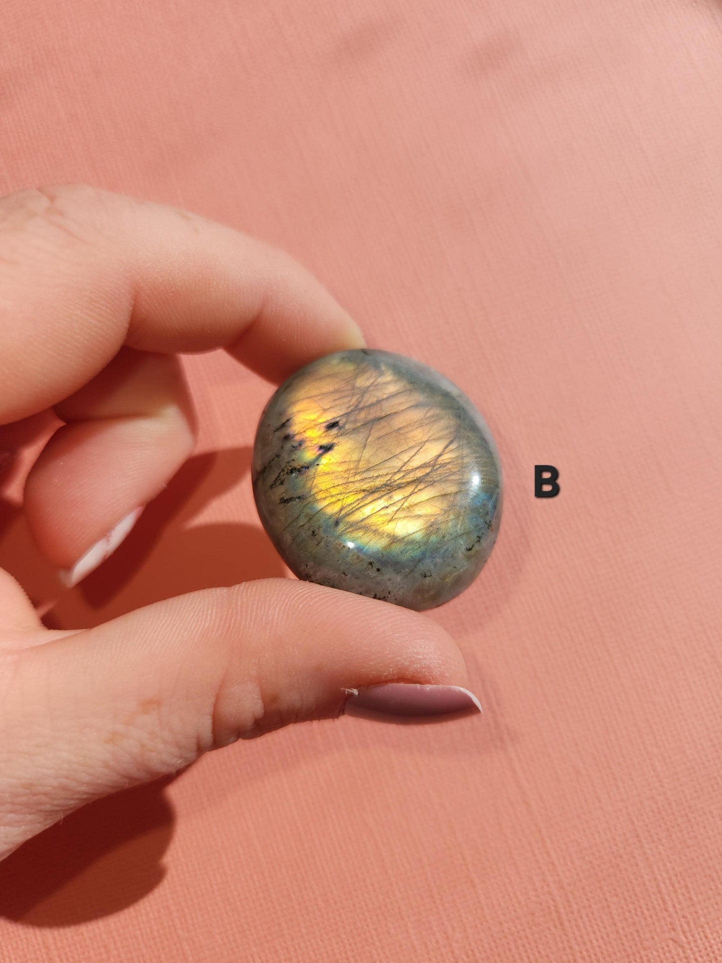 Detailed view of a Labradorite palm stone with a bright yellow flash on a pink background. Known for its powerful protective properties and luminous sheen, this polished gemstone supports transformation, energy shielding, and inner clarity. Great for meditation, chakra alignment, reiki practices, or as a unique gift for crystal enthusiasts and intuitive healers.