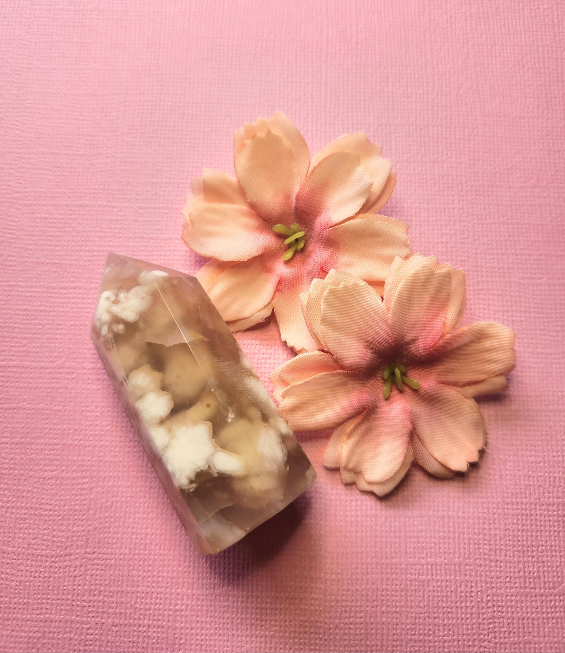Flower Agate crystal tower with soft pink and white patterns, displayed upright on a light pink background with two flower props — polished healing crystal point for manifestation, feminine energy, and emotional growth