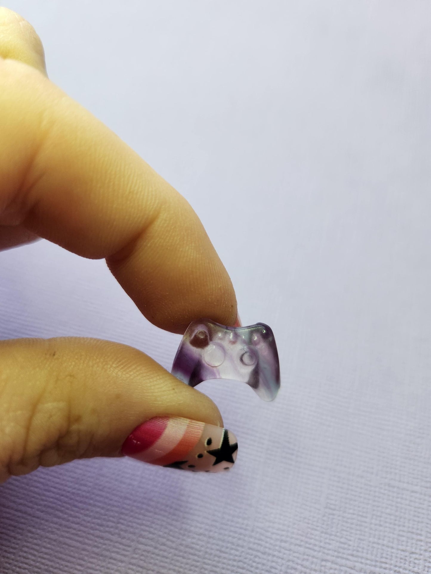 Close-up photo of a single fluorite mini game controller carving held between fingers, showing polished details and natural purple, green and blue fluorite banding against a lavender backdrop. Ideal for fluorite carvings, gamer-themed crystal collectibles, mini crystal decor and unique gemstone gifts