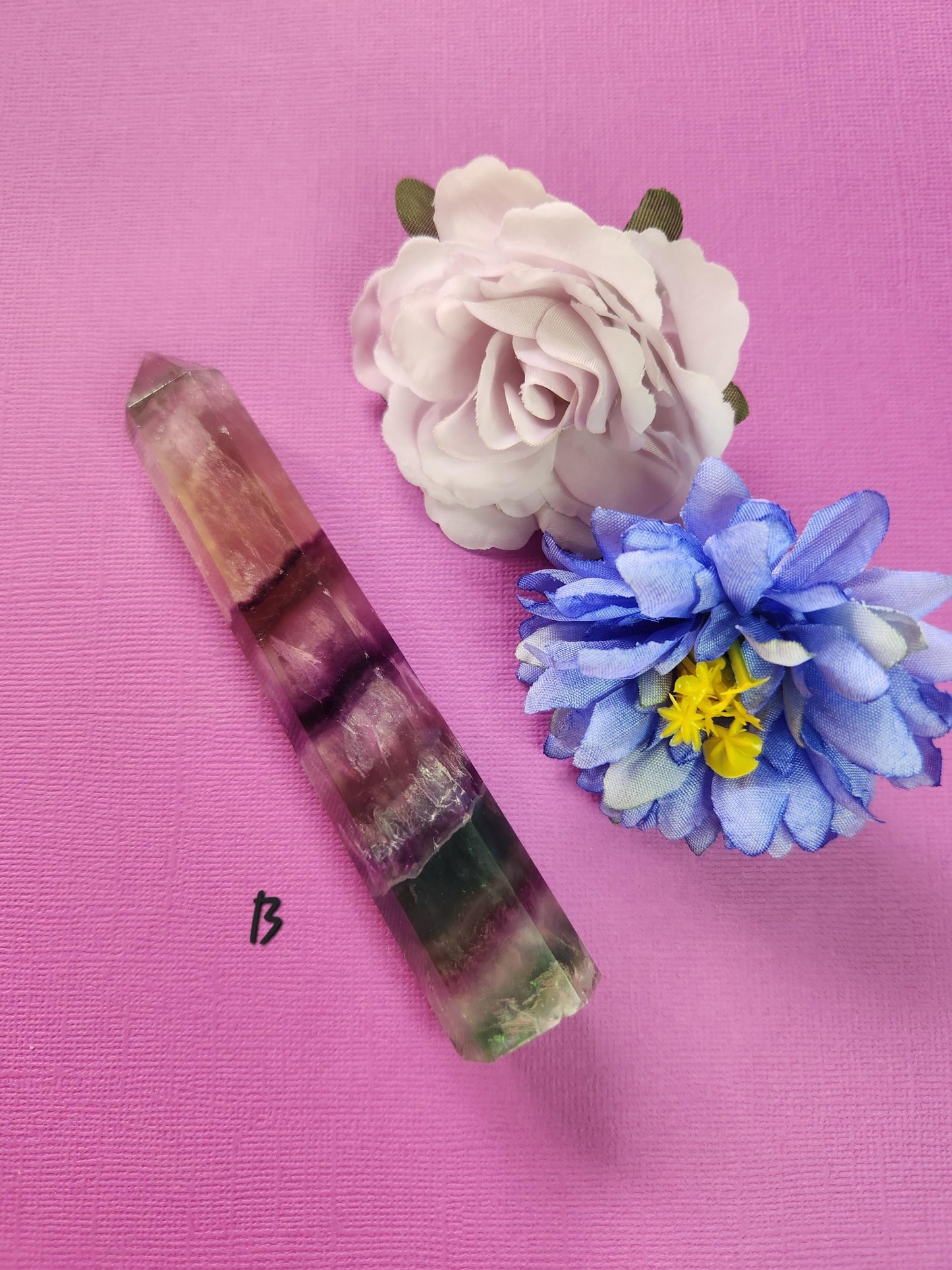 Polished Fluorite tower B showing unique green, purple, and translucent banding, styled with two flower props on a purple background, premium healing gemstone for clarity, meditation, chakra work, and metaphysical crystal décor