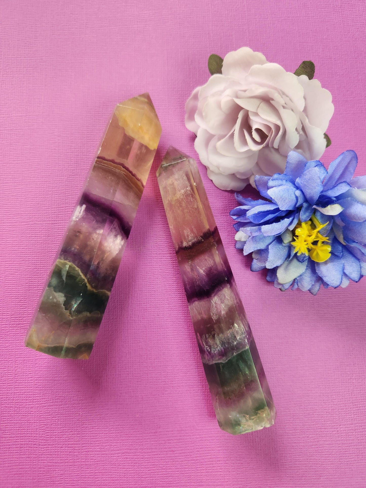 Two polished Fluorite crystal towers with natural green, purple, and clear banding, displayed with two flower props on a purple background, high-quality gemstone points for mental clarity, meditation, spiritual healing, chakra balancing, and metaphysical décor