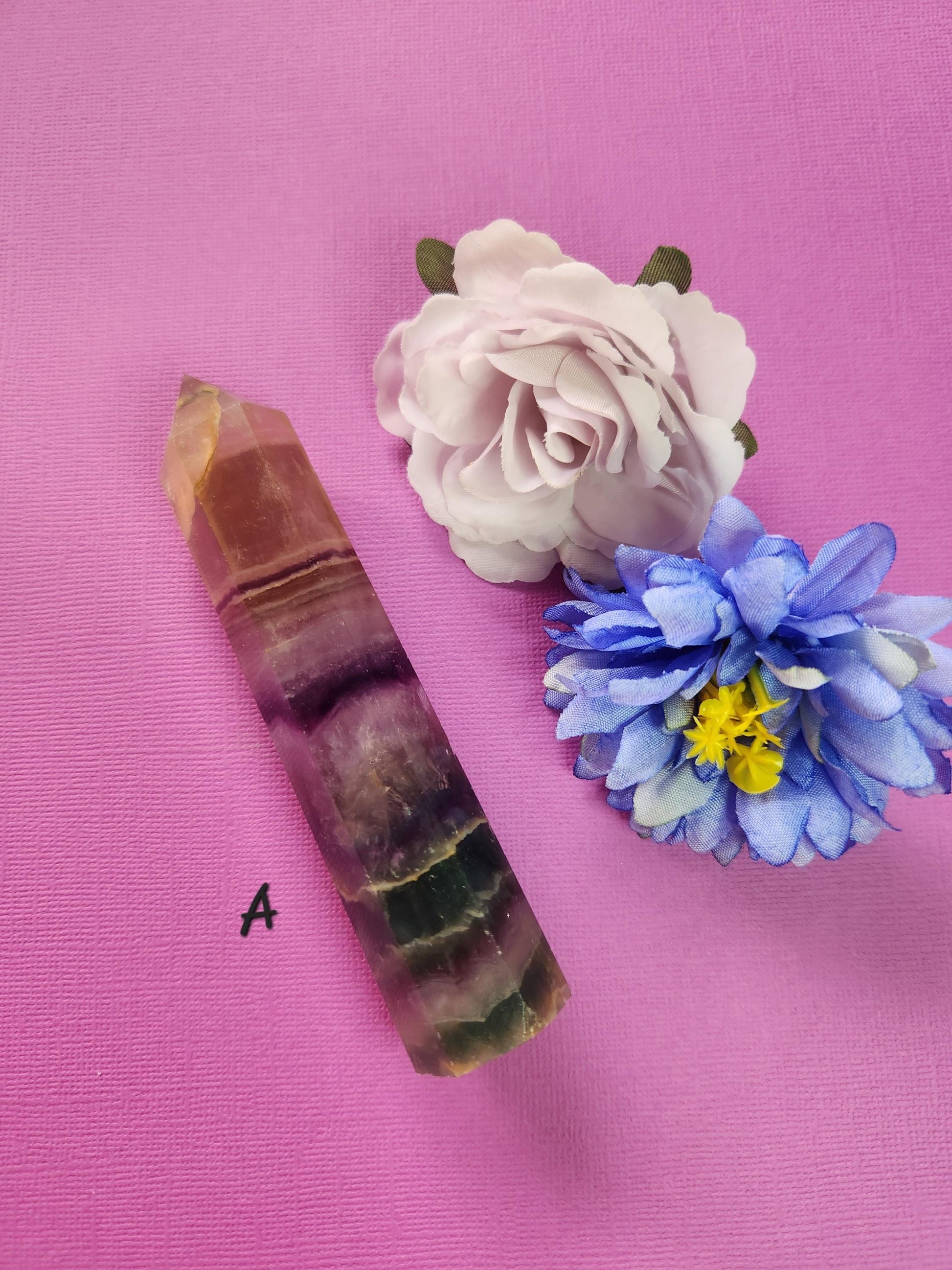 Polished Fluorite tower A with green, purple, and clear banding, displayed with two flower props on a purple background, natural crystal point for focus, energy cleansing, spiritual healing, meditation, and decorative crystal collection