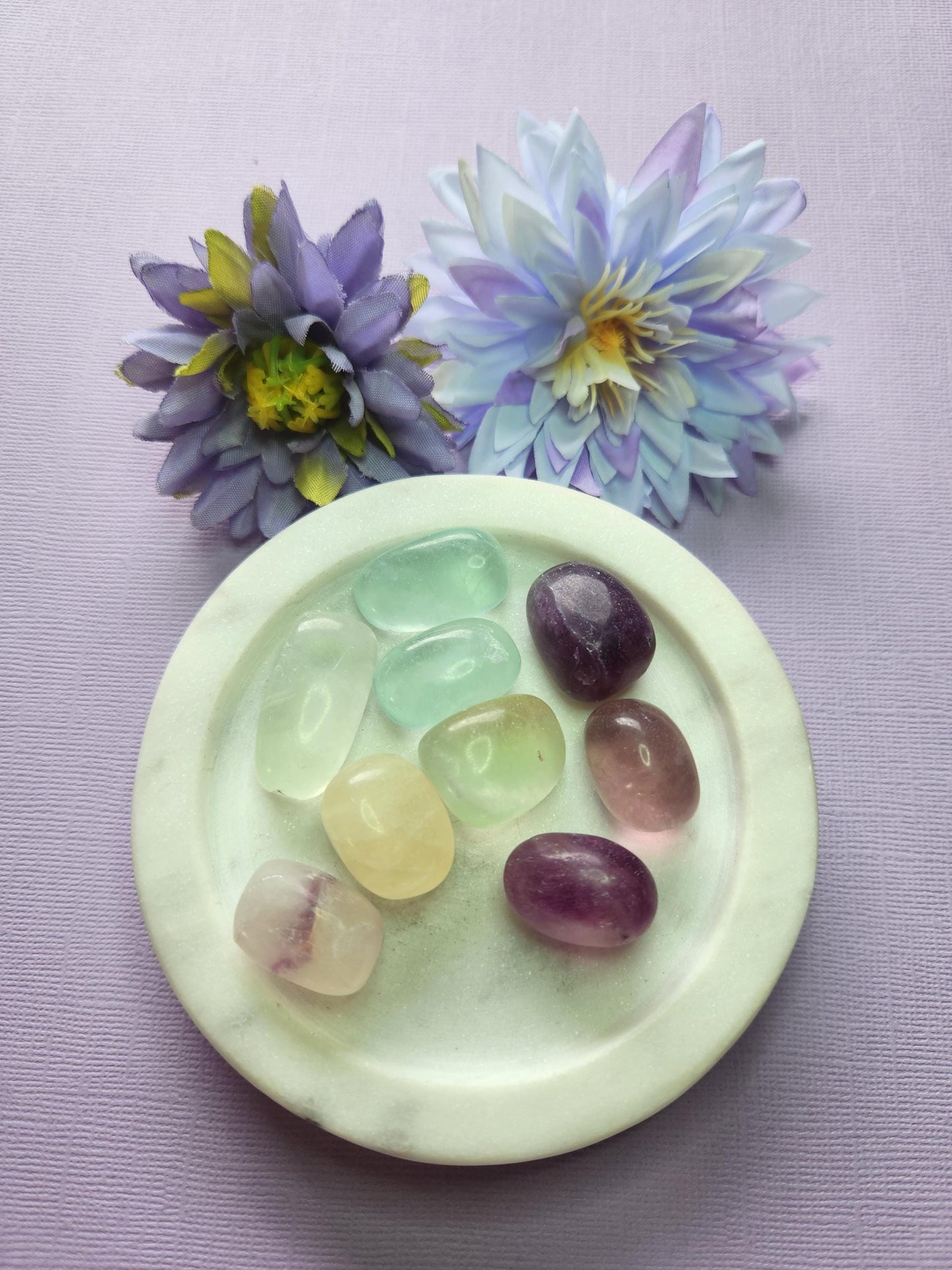 Close-up photo of several fluorite tumbles displayed in a white dish with two soft flower props on a lavender backdrop. The tumbles show natural purple, green and blue fluorite banding with a polished glossy finish. Ideal for fluorite crystals, tumbled gemstones, crystal collections, and decorative crystal accents.